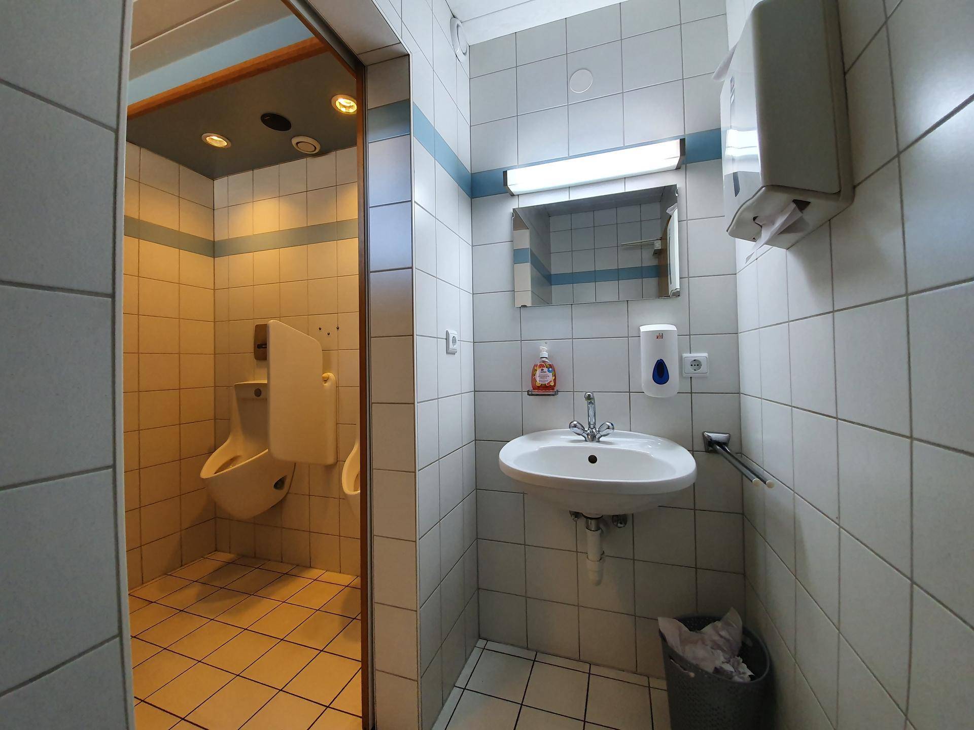 Sauberer Toilettenraum mit Waschbecken, Spiegel und Urinal, gefliest in Weiß und Blau.