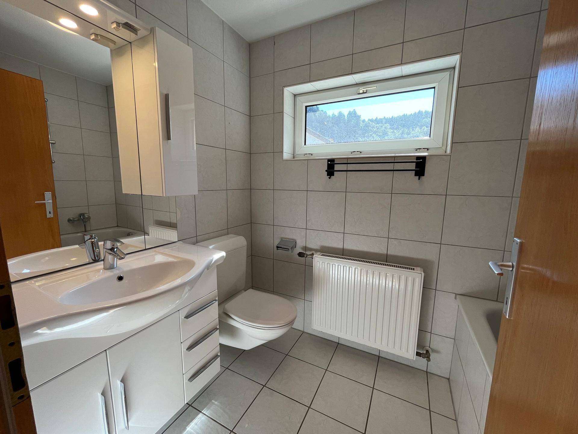 Helles Badezimmer mit Waschbecken, WC und Badewanne, gefliest in Weiß und Grau.