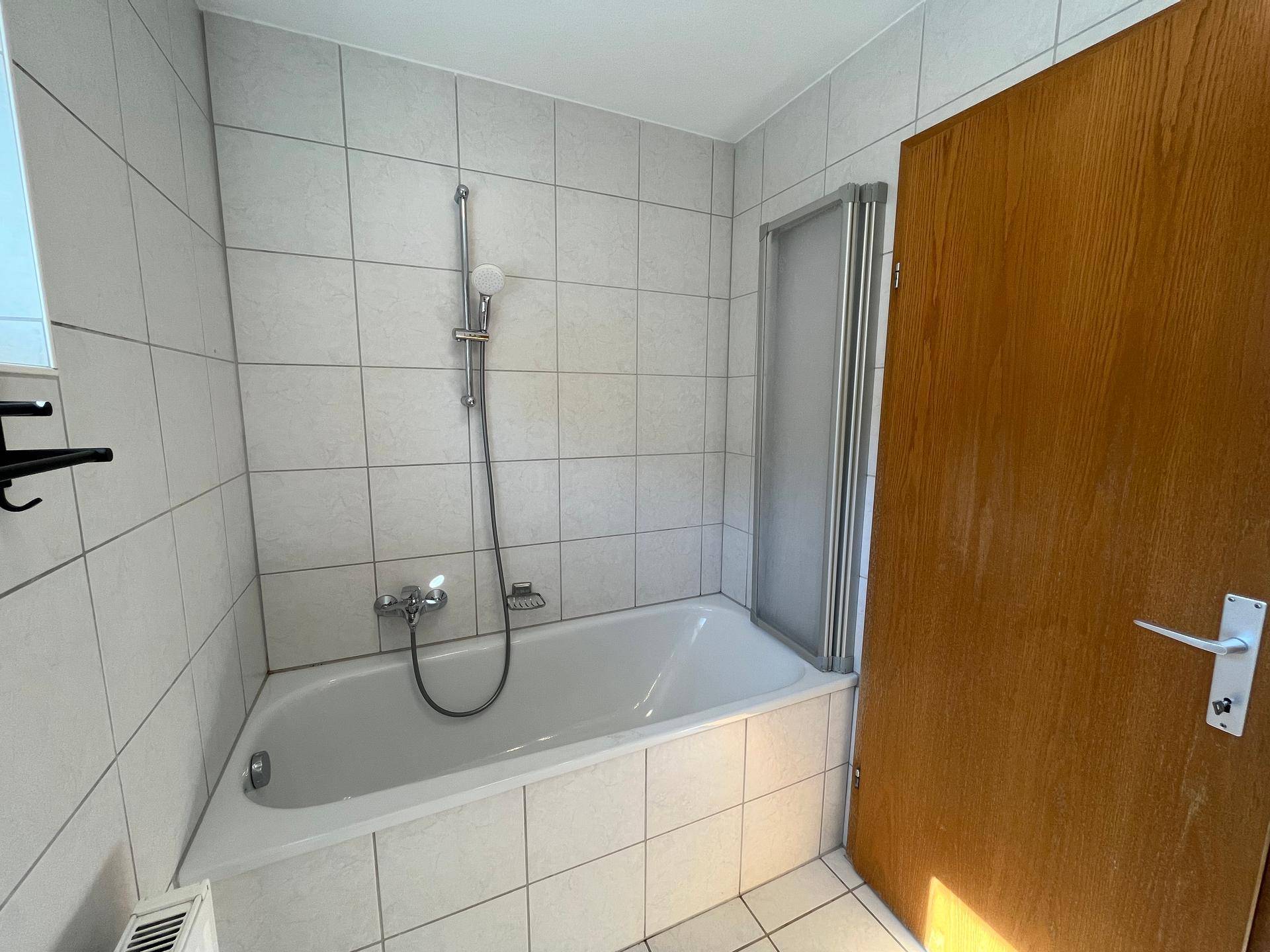 Badezimmer mit Badewanne und Duschabtrennung, gefliesten Wänden und Holztür.