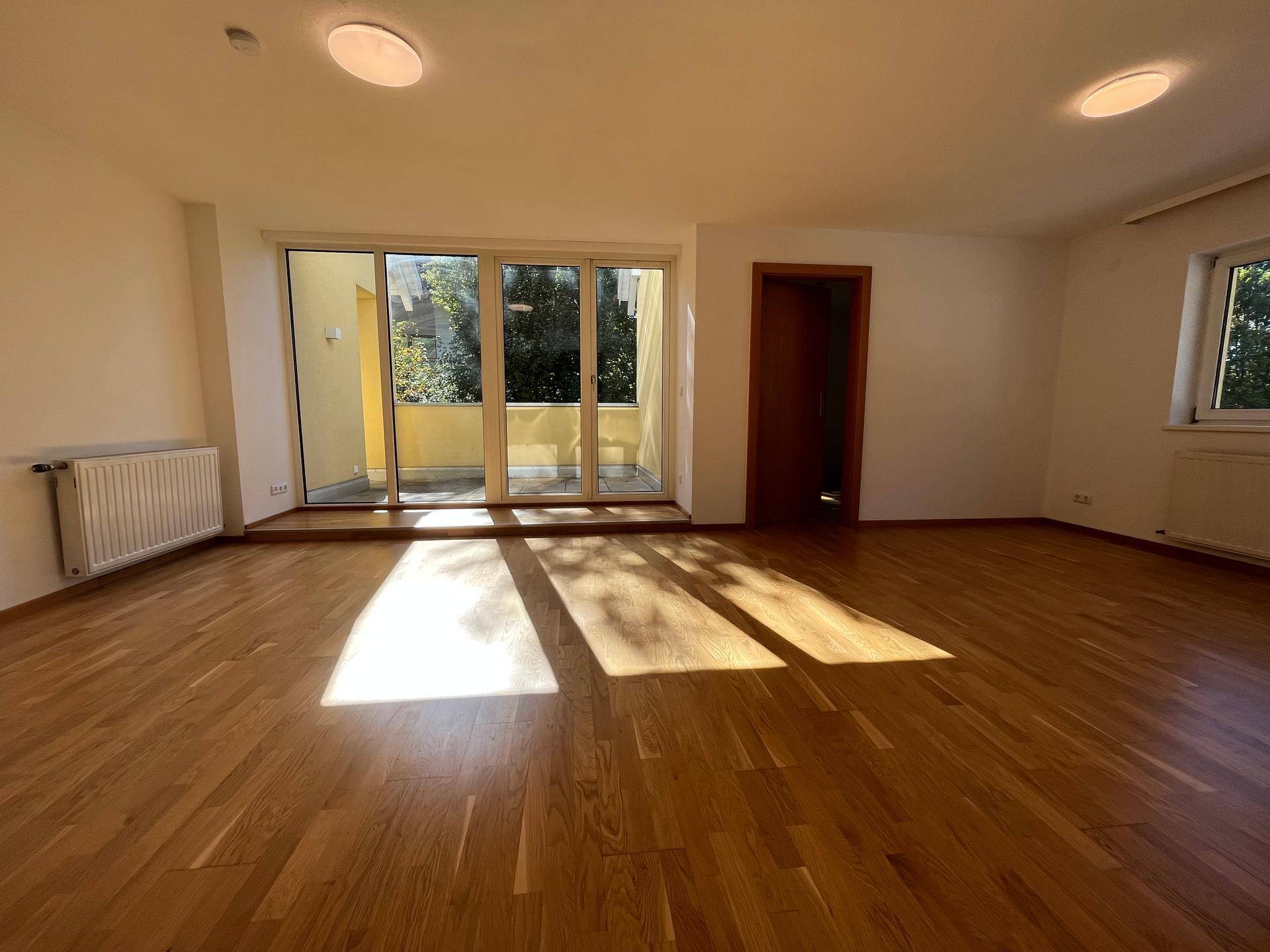 Heller Wohnbereich mit Holzboden und großen Fenstern, die viel Sonnenlicht hereinlassen.