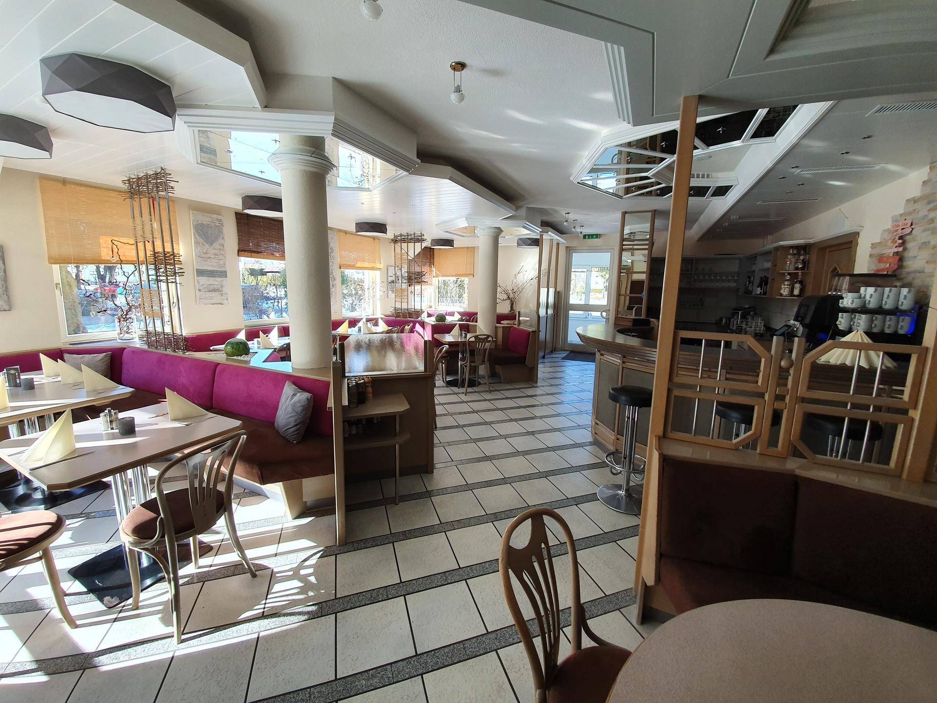 Weitläufiger Restaurantraum mit verschiedenen Sitzbereichen, Bar und Fliesenboden, gut beleuchtet.