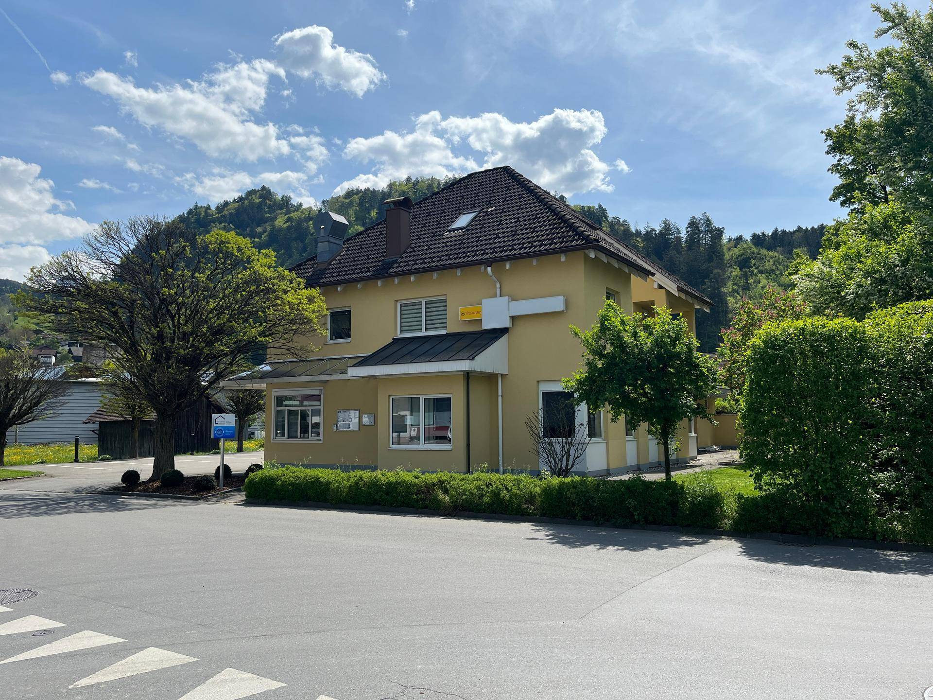 Gelbes Gebäude mit Satteldach, umgeben von Bäumen und Grünflächen unter blauem Himmel.