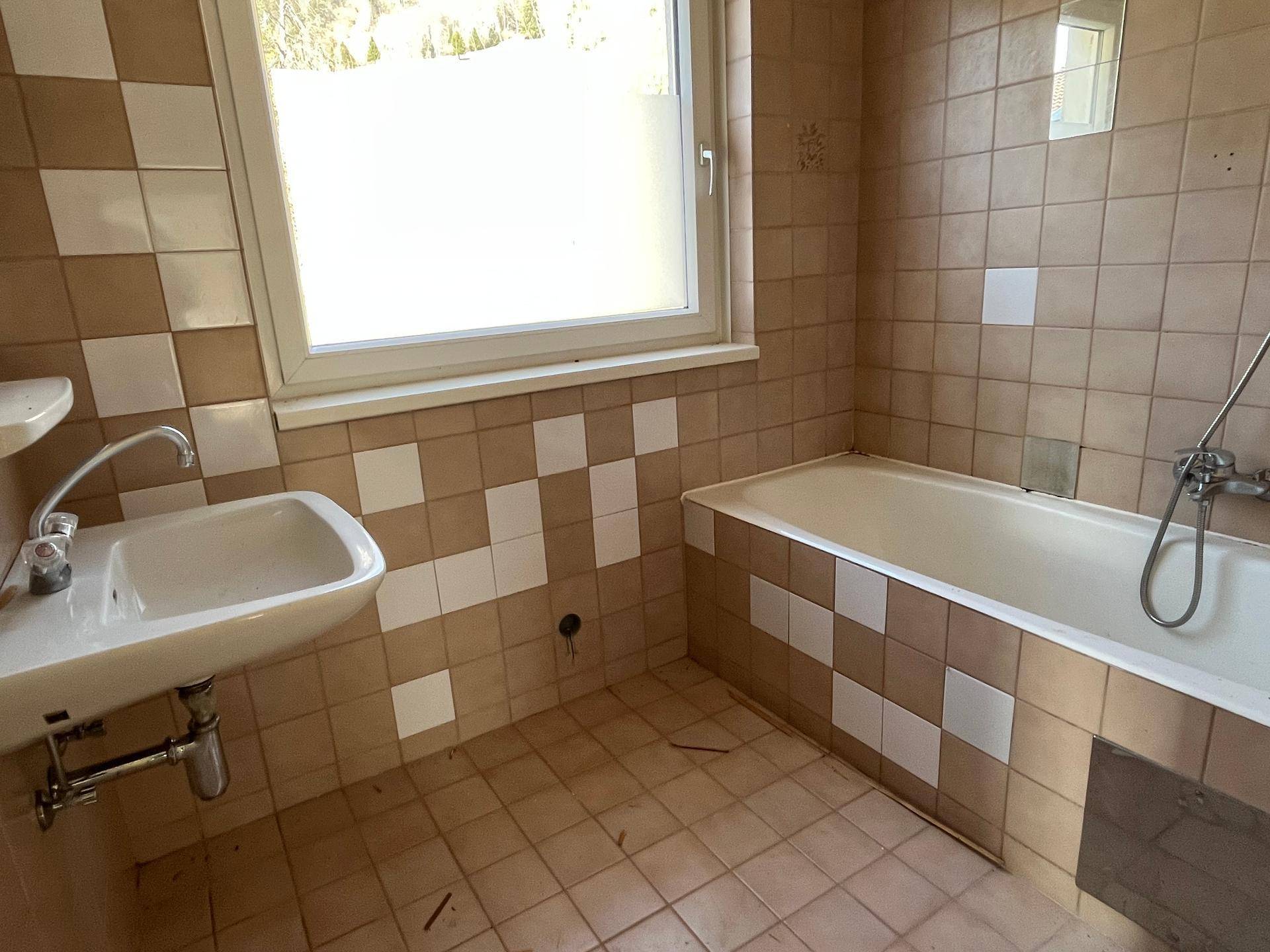 Badezimmer mit Badewanne, Waschbecken und Fenster, das eine umfassende Renovierung erfordert.