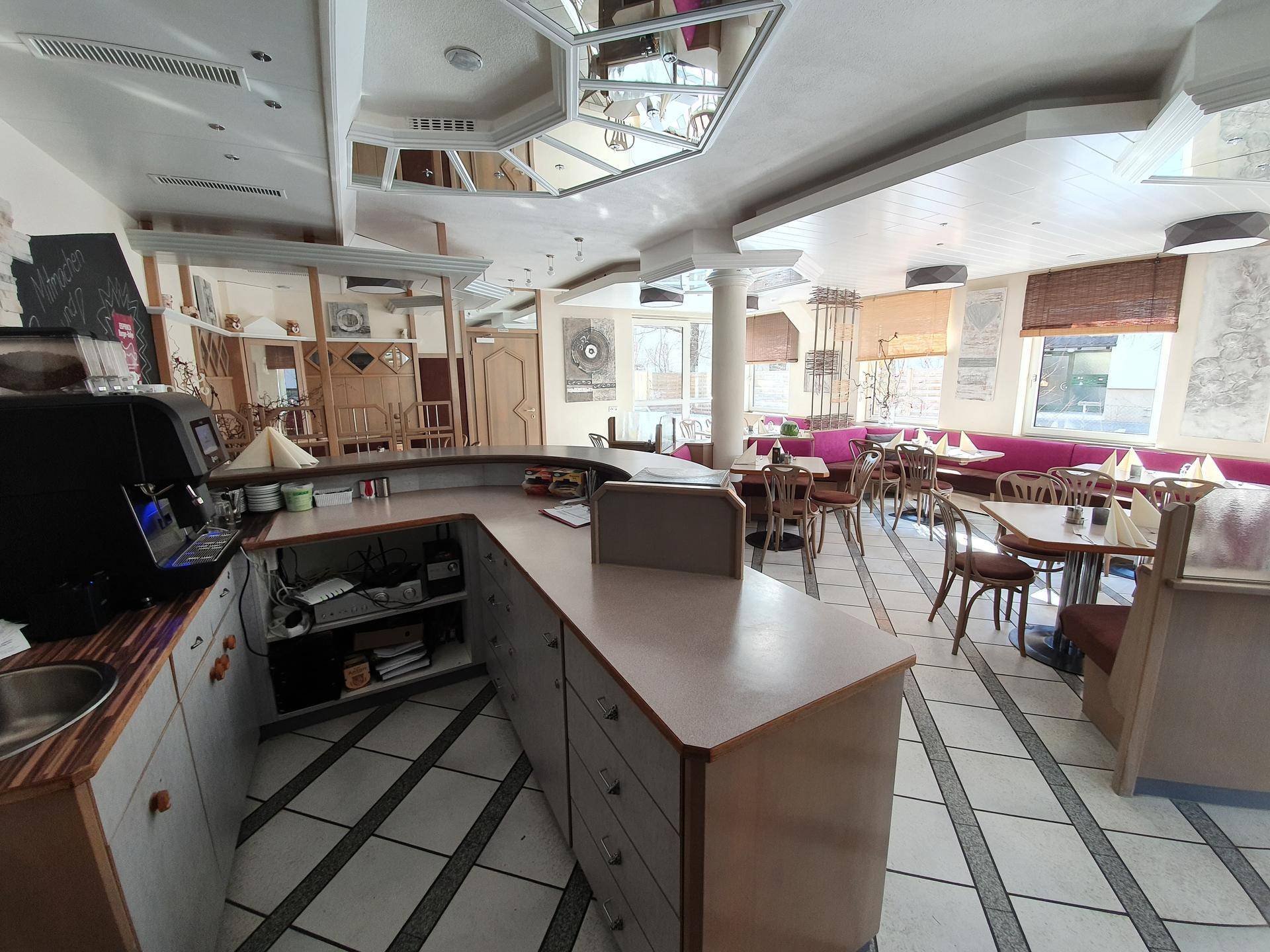 Innenansicht eines Restaurants mit Theke, Sitzbänken und Tischen, beleuchtet durch natürliches Licht.