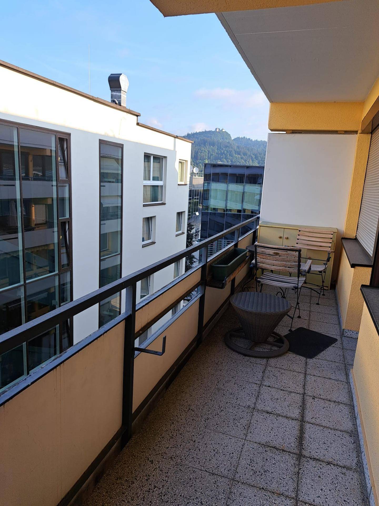 Sonnenverwöhnter Balkon mit Fliesenboden, Sitzgelegenheiten und Blick auf die umliegenden Gebäude und Hügel.