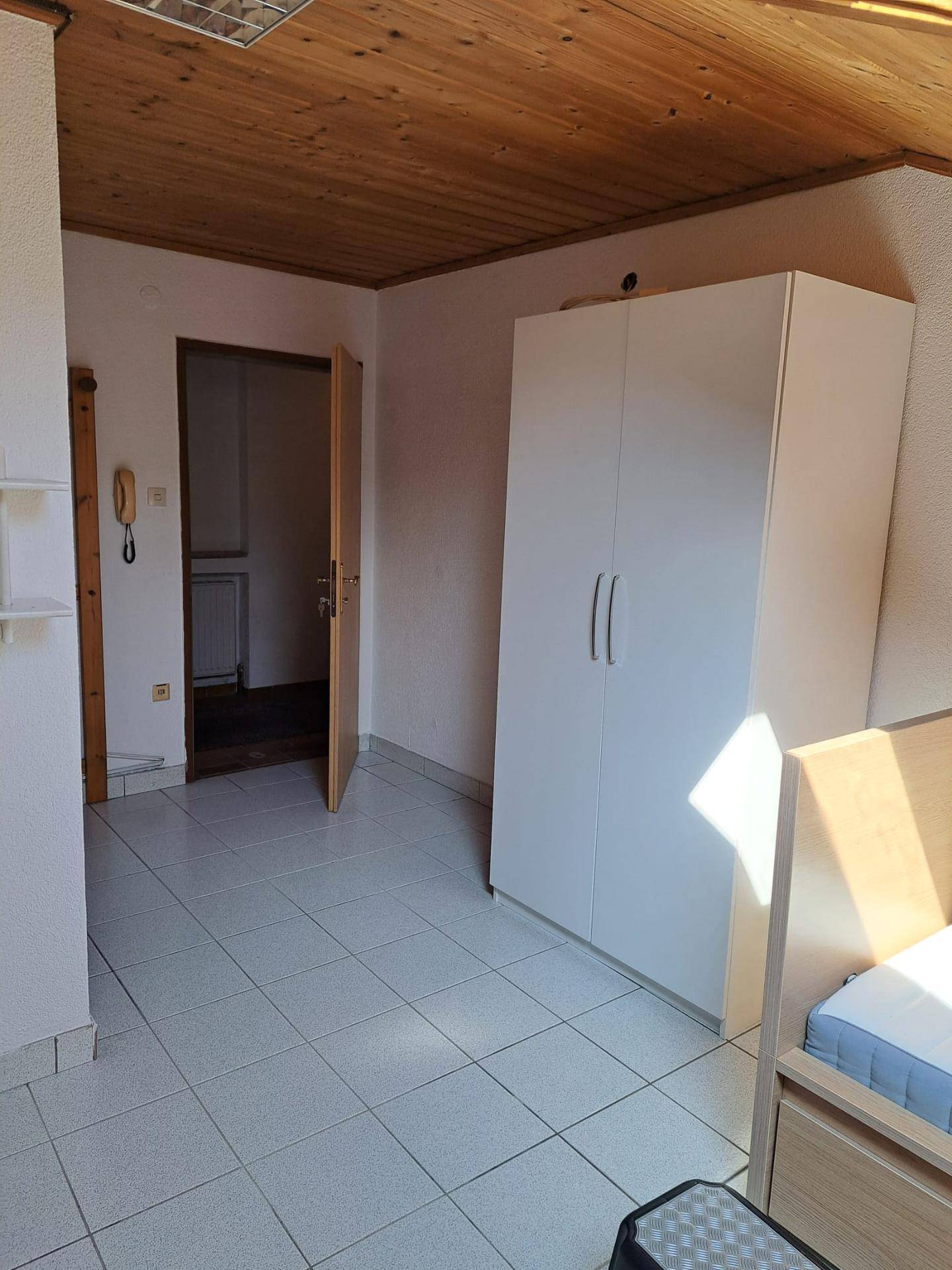 Schlafzimmer mit Fliesenboden, Holzdecke und weißem Kleiderschrank, mit Zugang zu einem weiteren Raum.