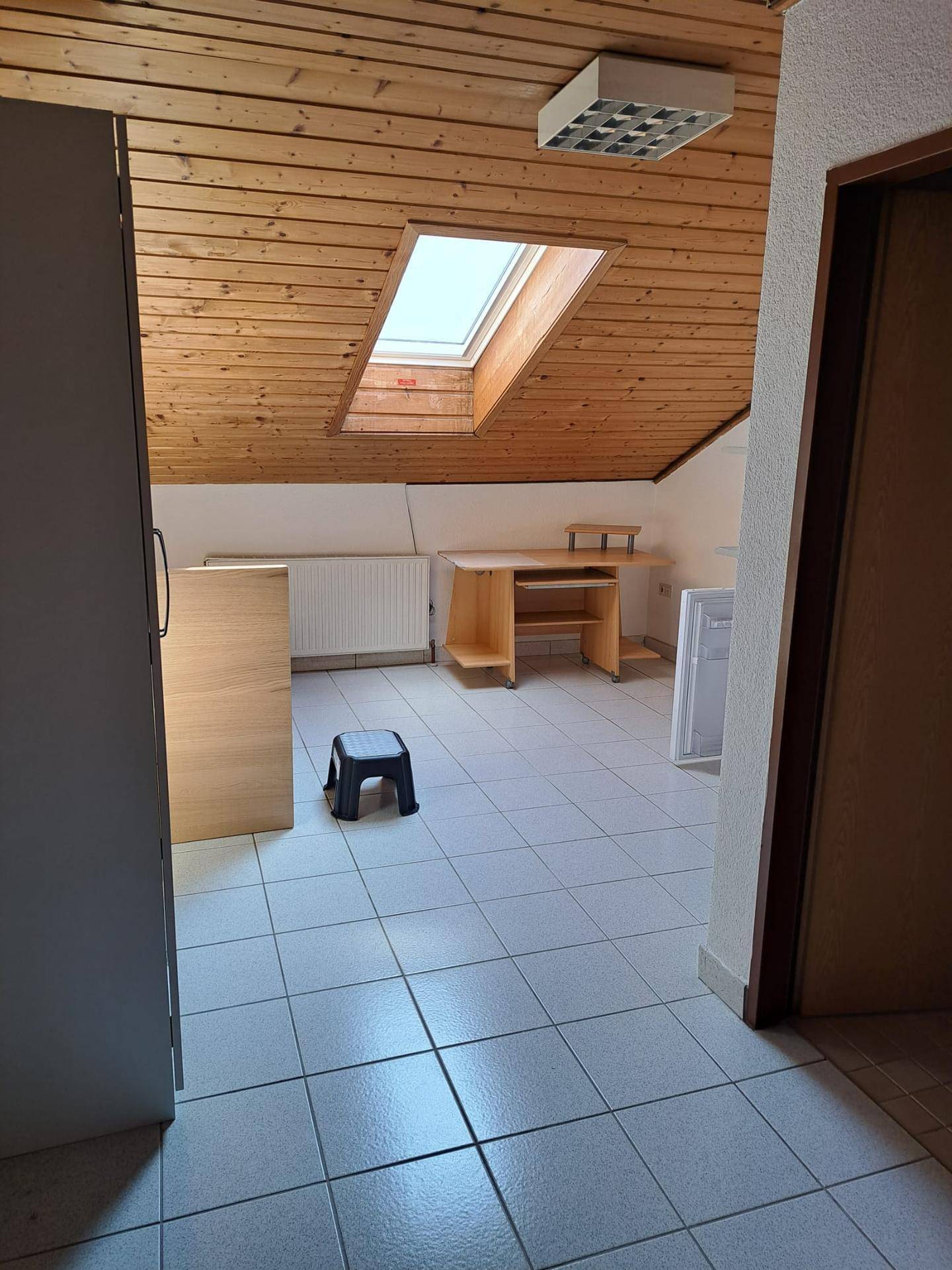Dachzimmer mit Holzdecke, Dachfenster und Fliesenboden, ausgestattet mit einem Schreibtisch.