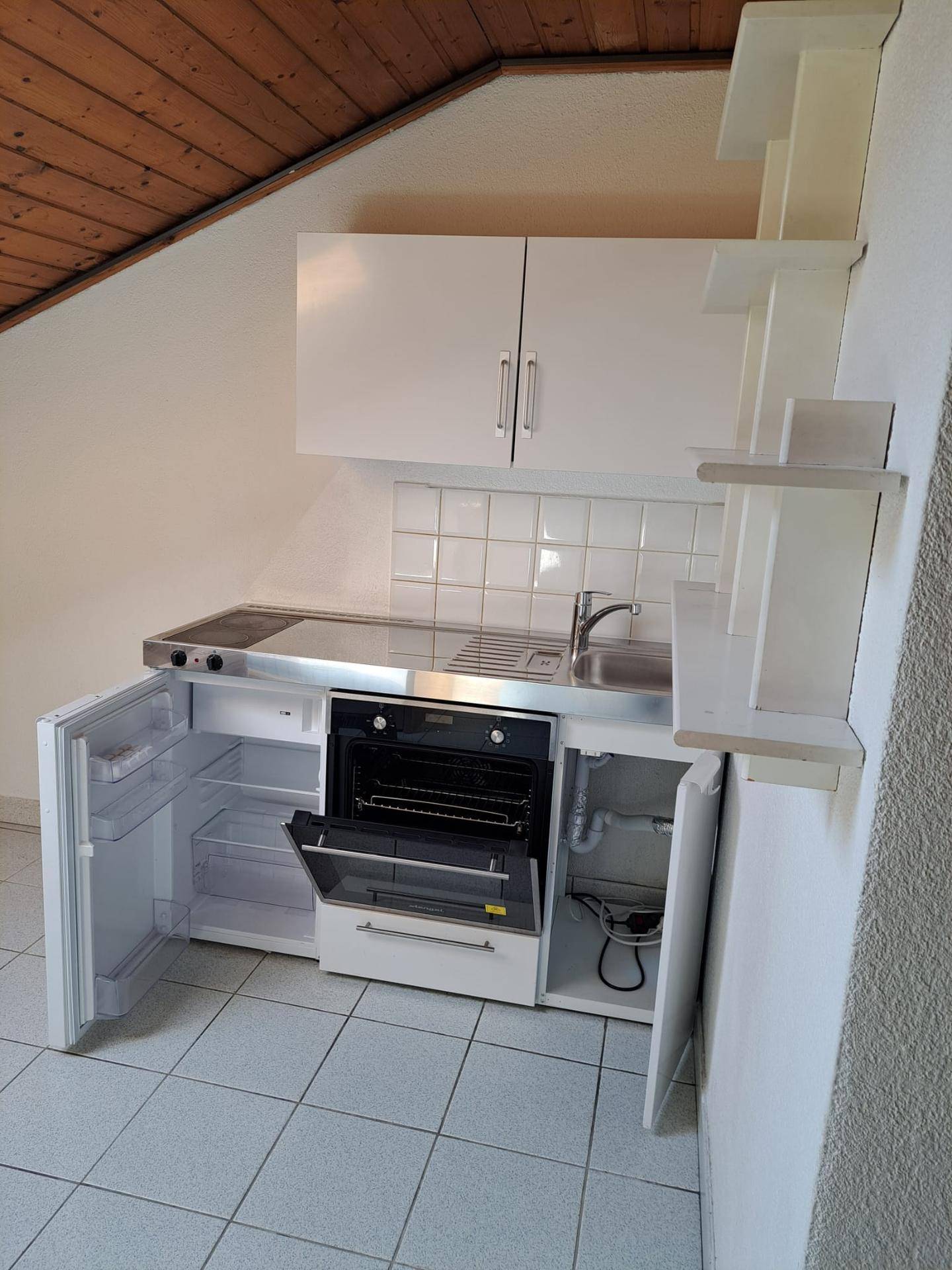 Kompakte Küchenzeile unter einer Holzschräge mit integrierten Geräten und Fliesenboden.