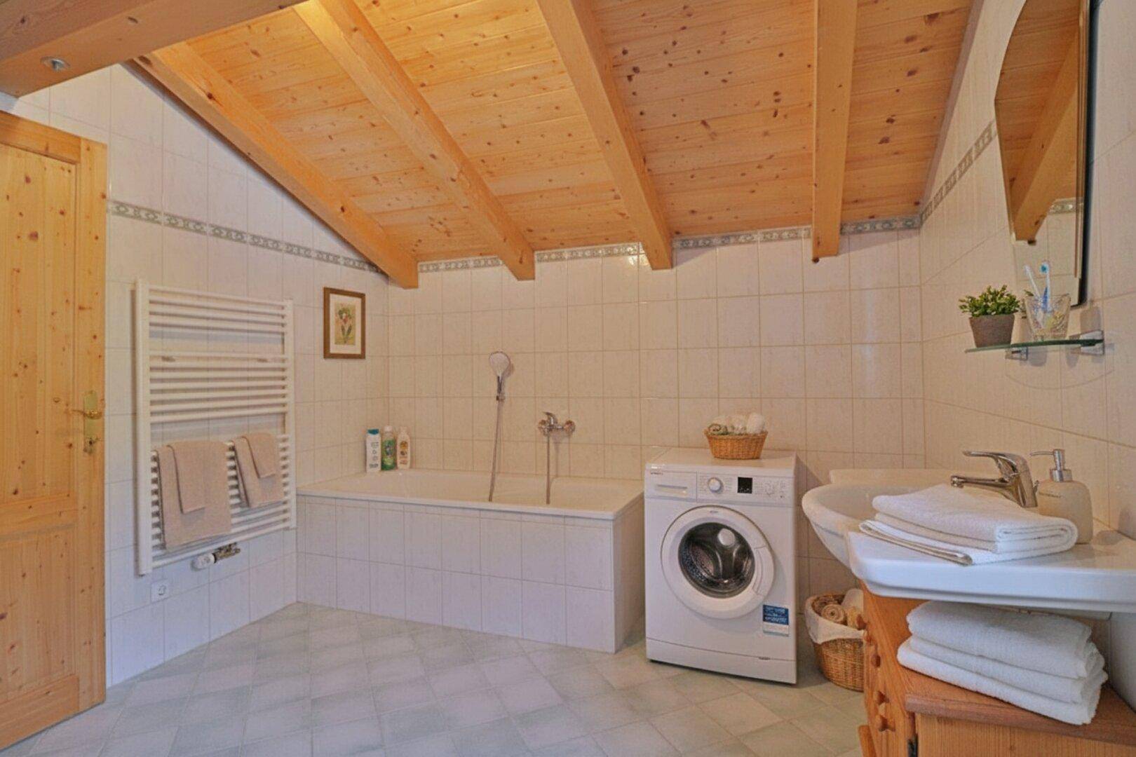 Gefliestes Badezimmer mit Badewanne, Waschmaschine und Dachschräge, hell und funktional.