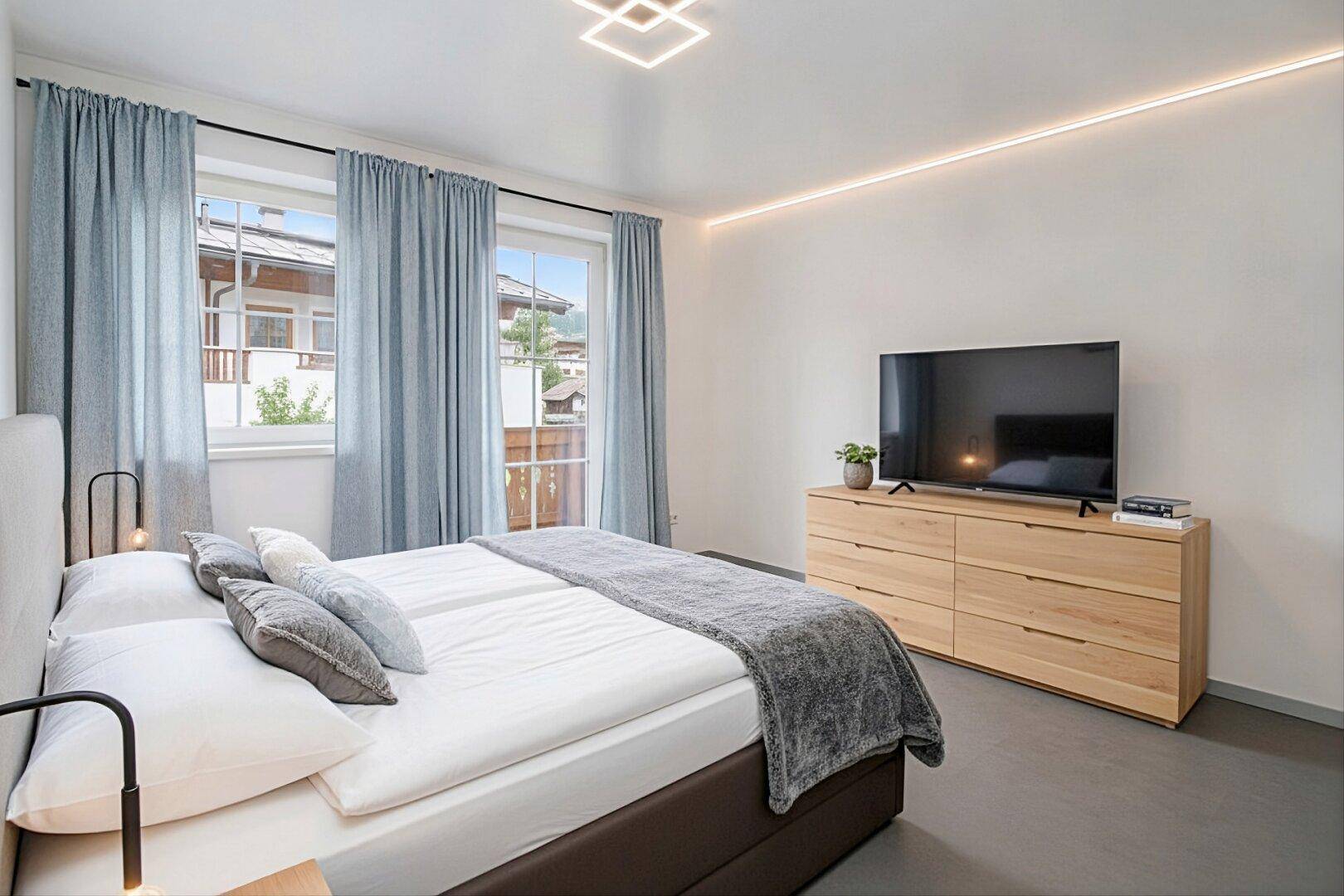Modernes Schlafzimmer mit Doppelbett, Holzkommode und großen Fenstern für viel Tageslicht.