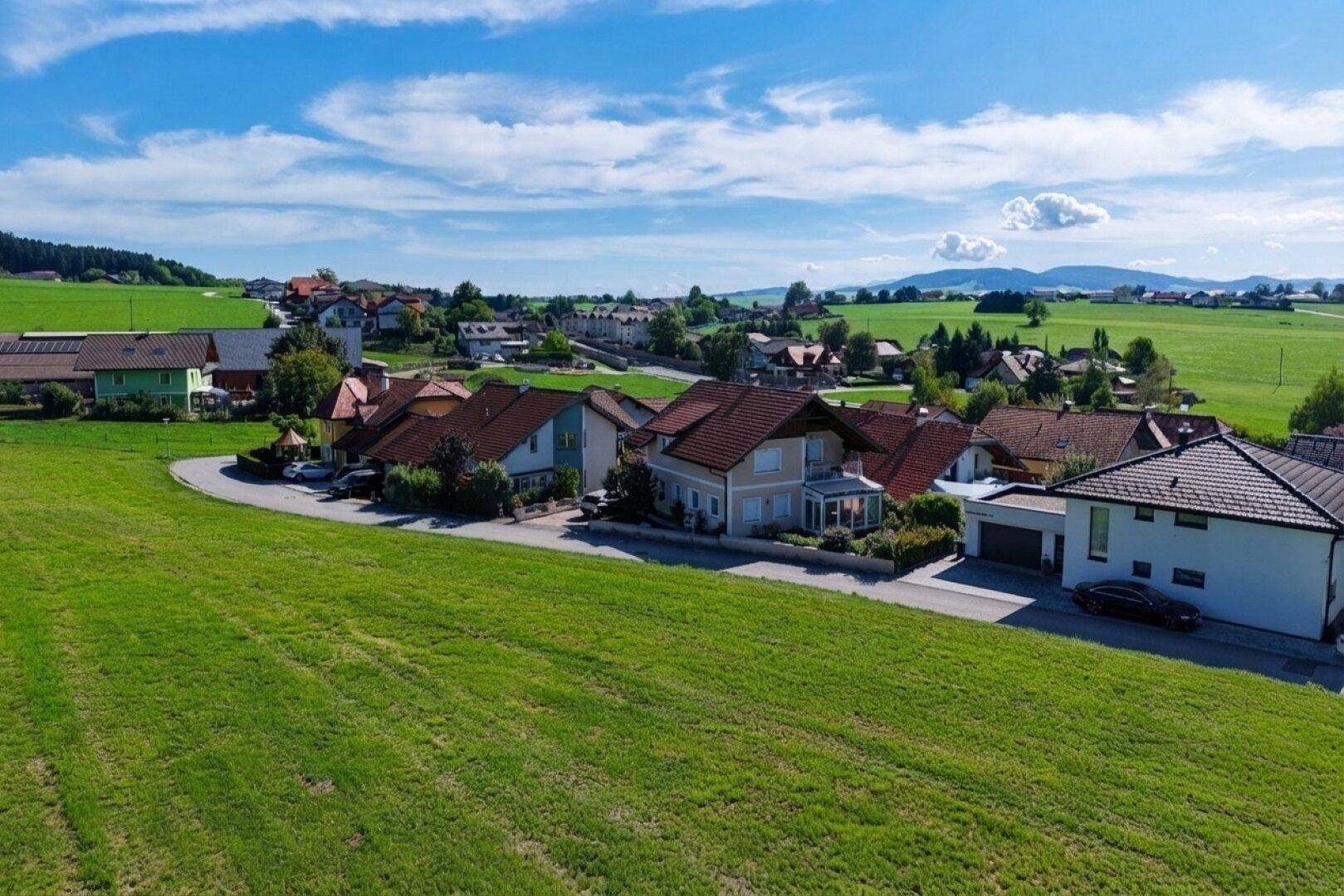 Panoramablick auf eine idyllische Wohngegend, umgeben von weiten Wiesen und einer Bergkette am Horizont.