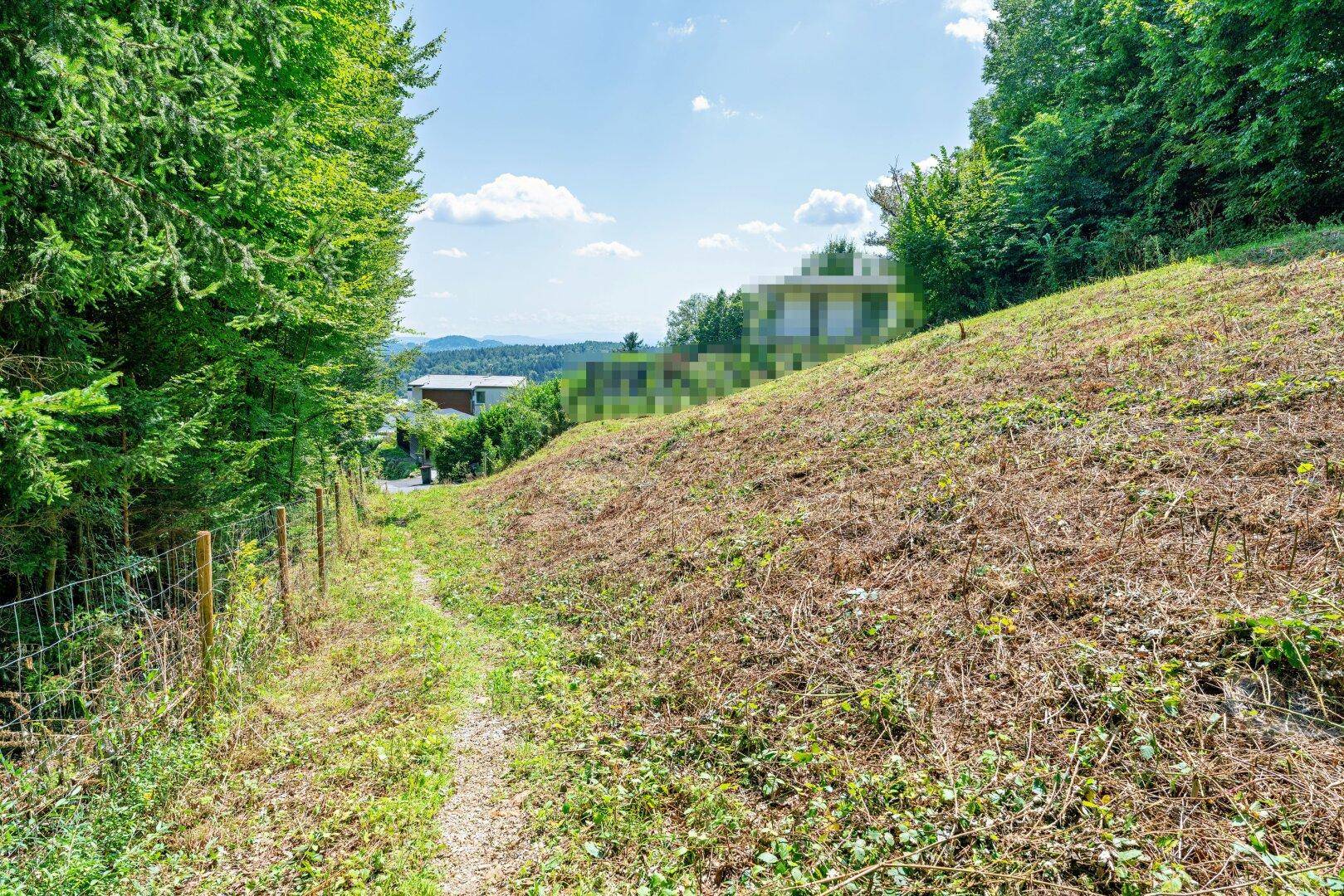 Weg führt einen grasbewachsenen Hang hinauf mit Blick auf entfernte Gebäude und Hügel.