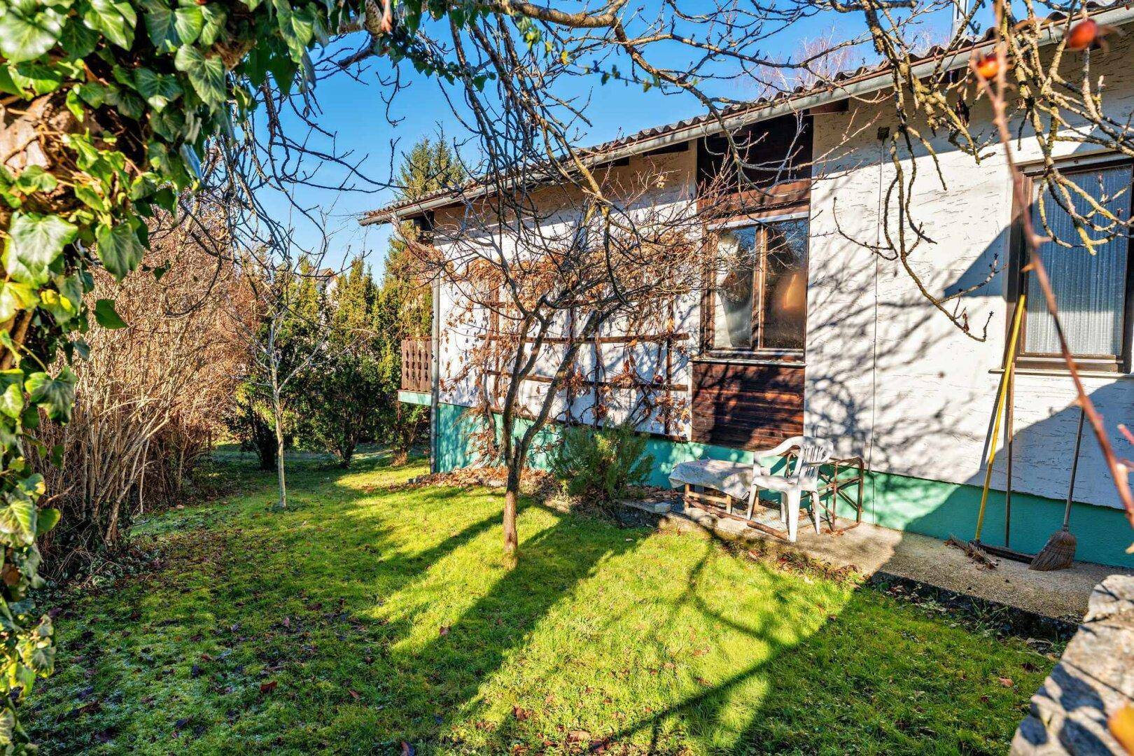 Seitenansicht des Hauses mit grünem Garten, Bäumen und einer kleinen Terrasse im Sonnenlicht.