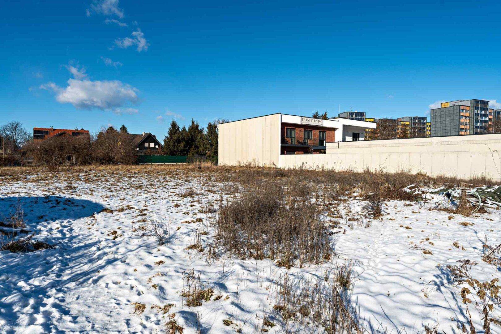 Offenes Feld mit Schneeresten und trockener Vegetation, im Hintergrund moderne Gebäude unter blauem Himmel.