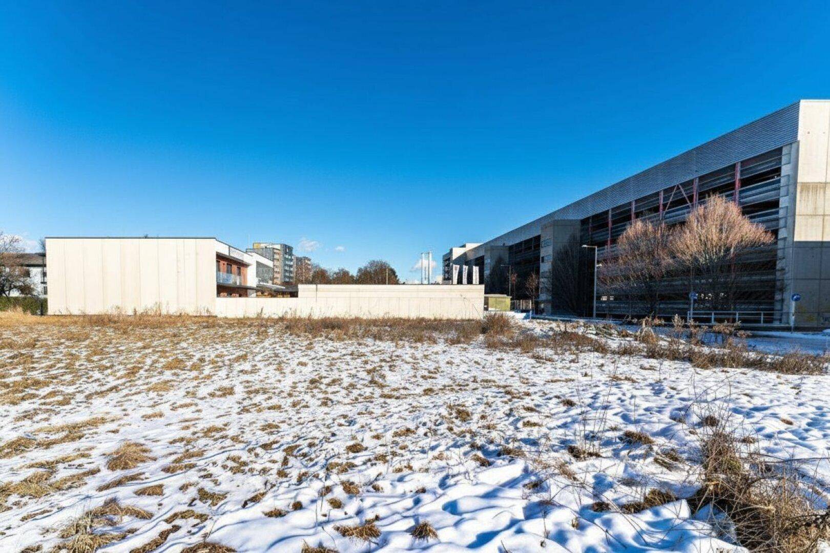Offenes Feld mit Schneeresten und trockener Vegetation, im Hintergrund große moderne Gebäude unter blauem Himmel.