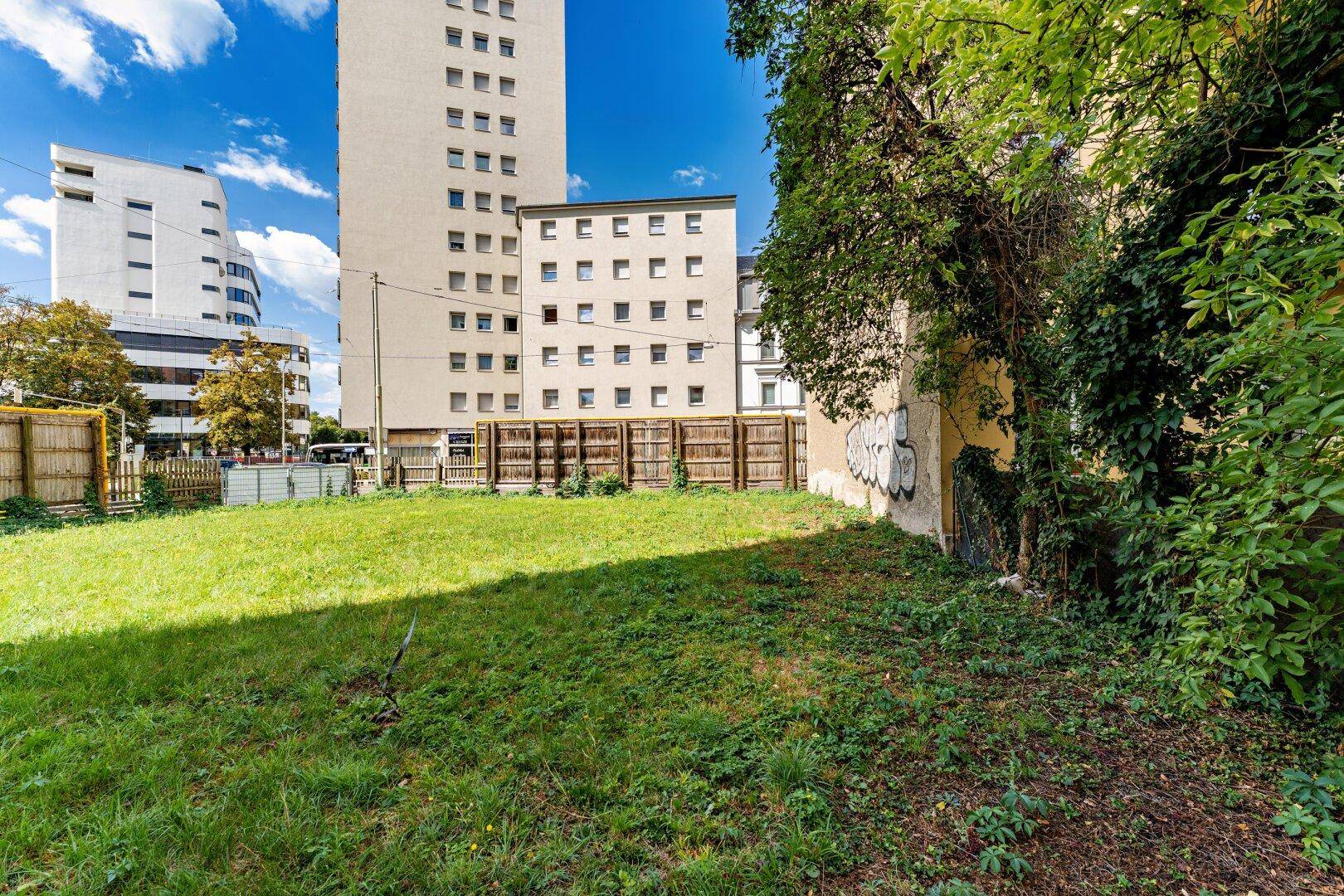 Unbebautes Grundstück mit Gras und Zäunen, im Hintergrund hohe Gebäude der städtischen Skyline.