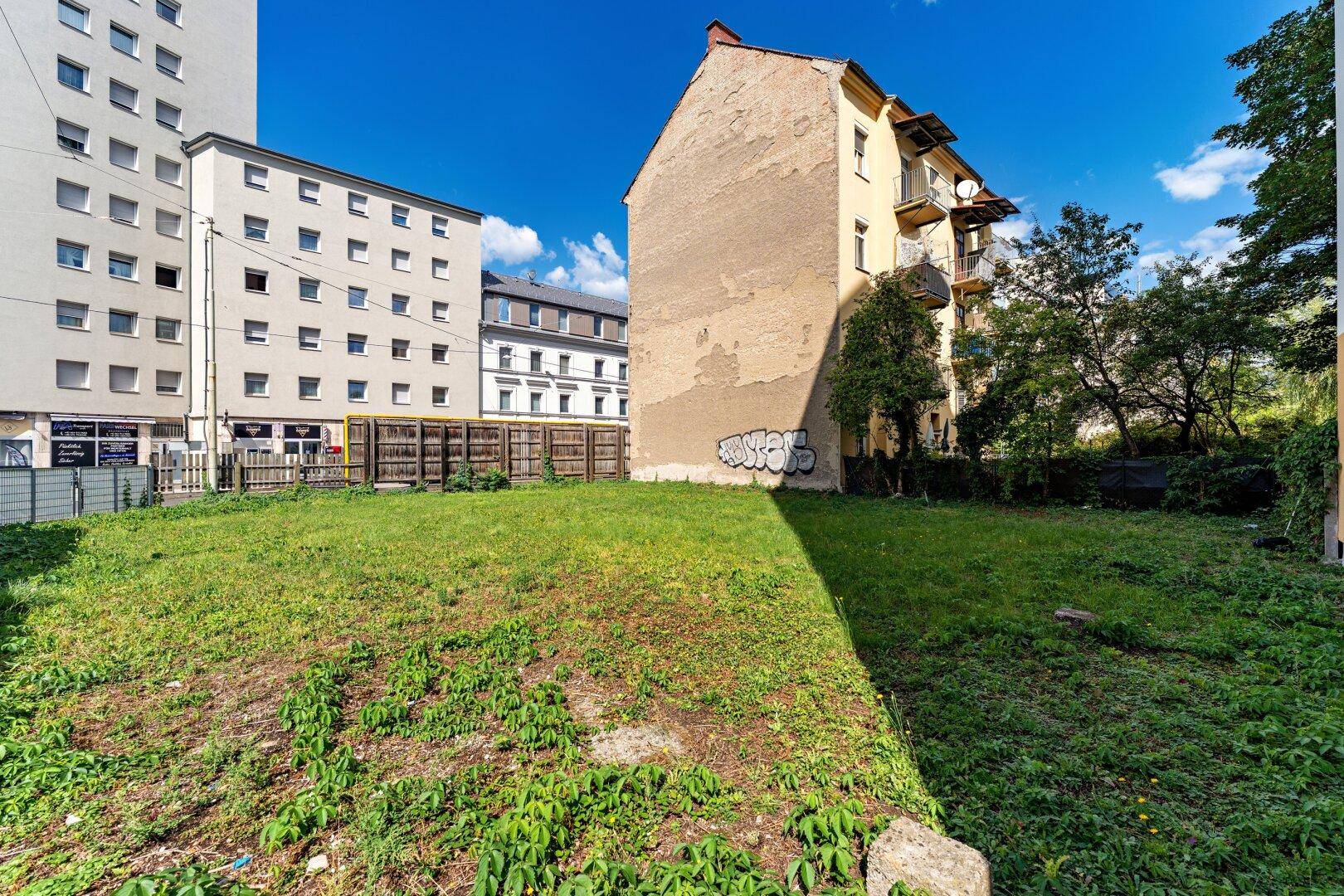 Grüne Grasfläche des unbebauten Grundstücks, umgeben von städtischen Gebäuden und einem blauen Himmel.