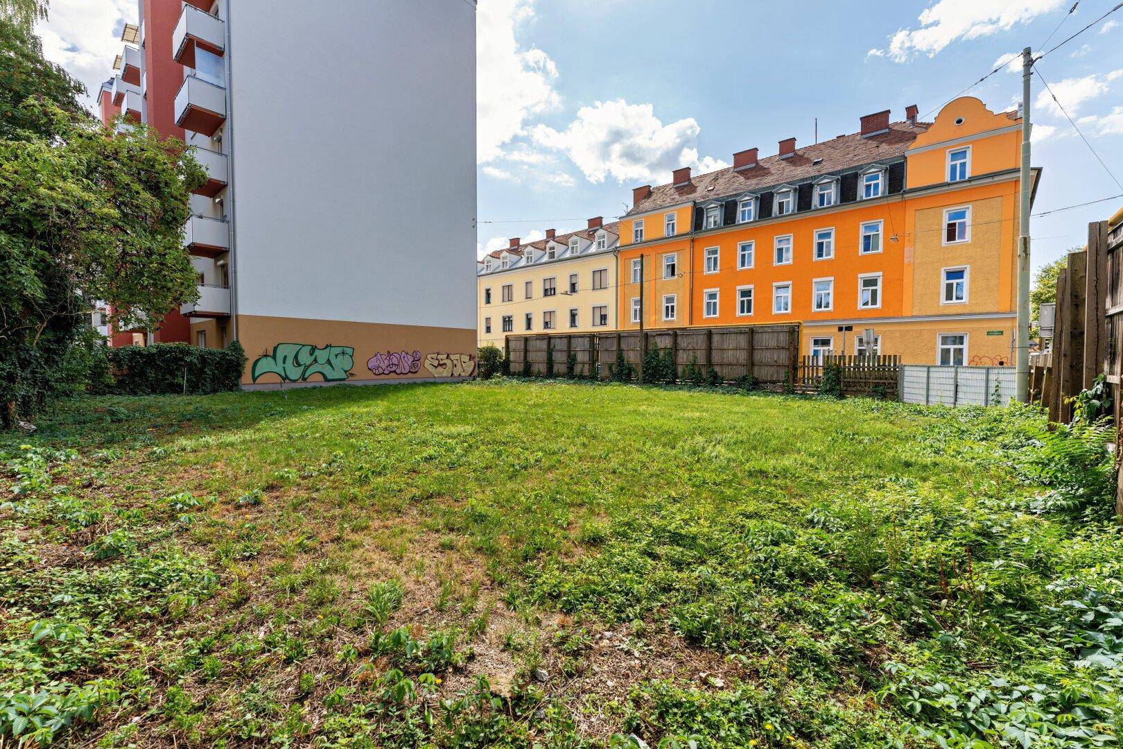 Blick auf das unbebaute Grundstück mit Grasfläche und umliegenden Wohngebäuden unter blauem Himmel.