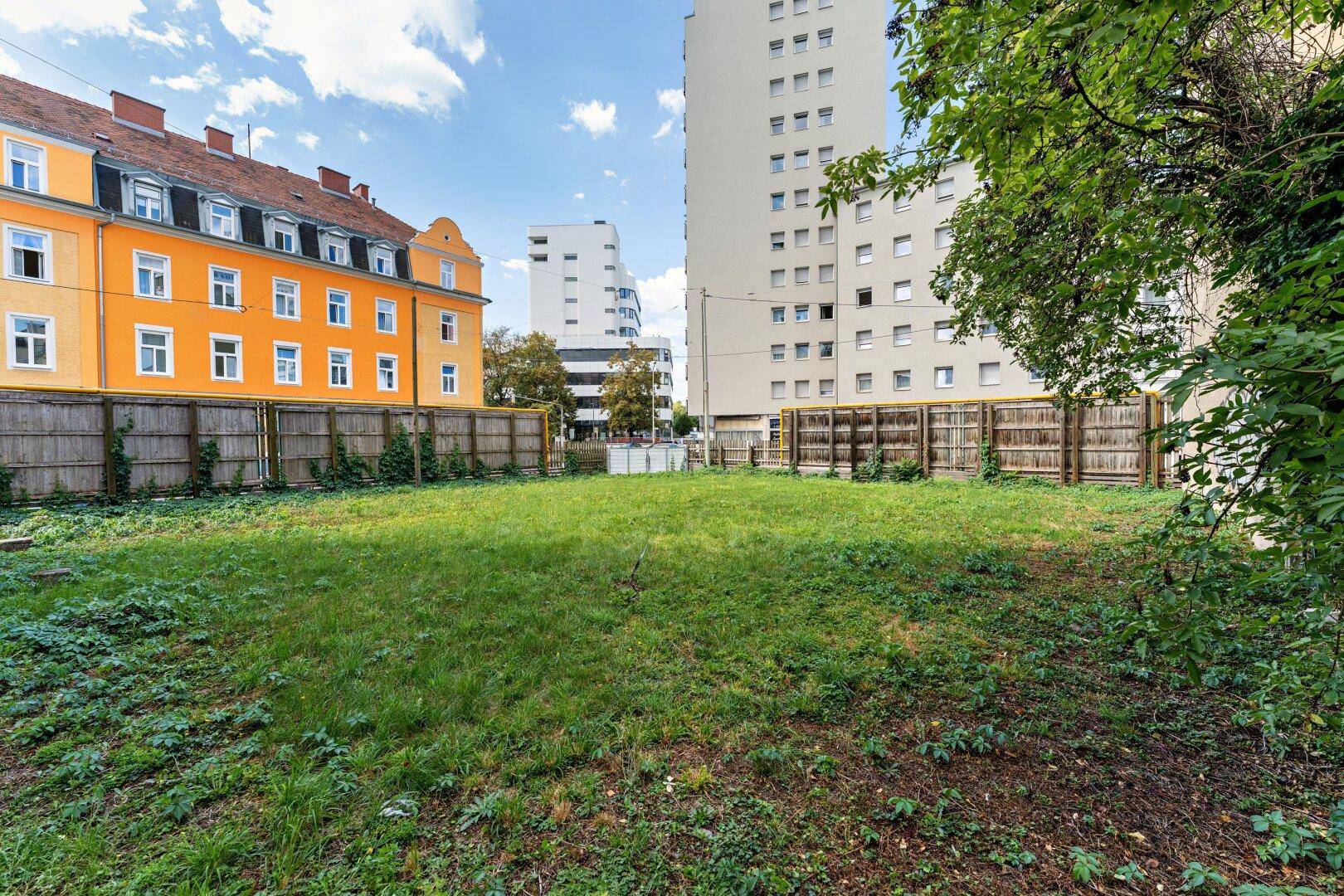 Weitläufige Grasfläche des unbebauten Grundstücks, umgeben von Holzzäunen und städtischen Gebäuden.