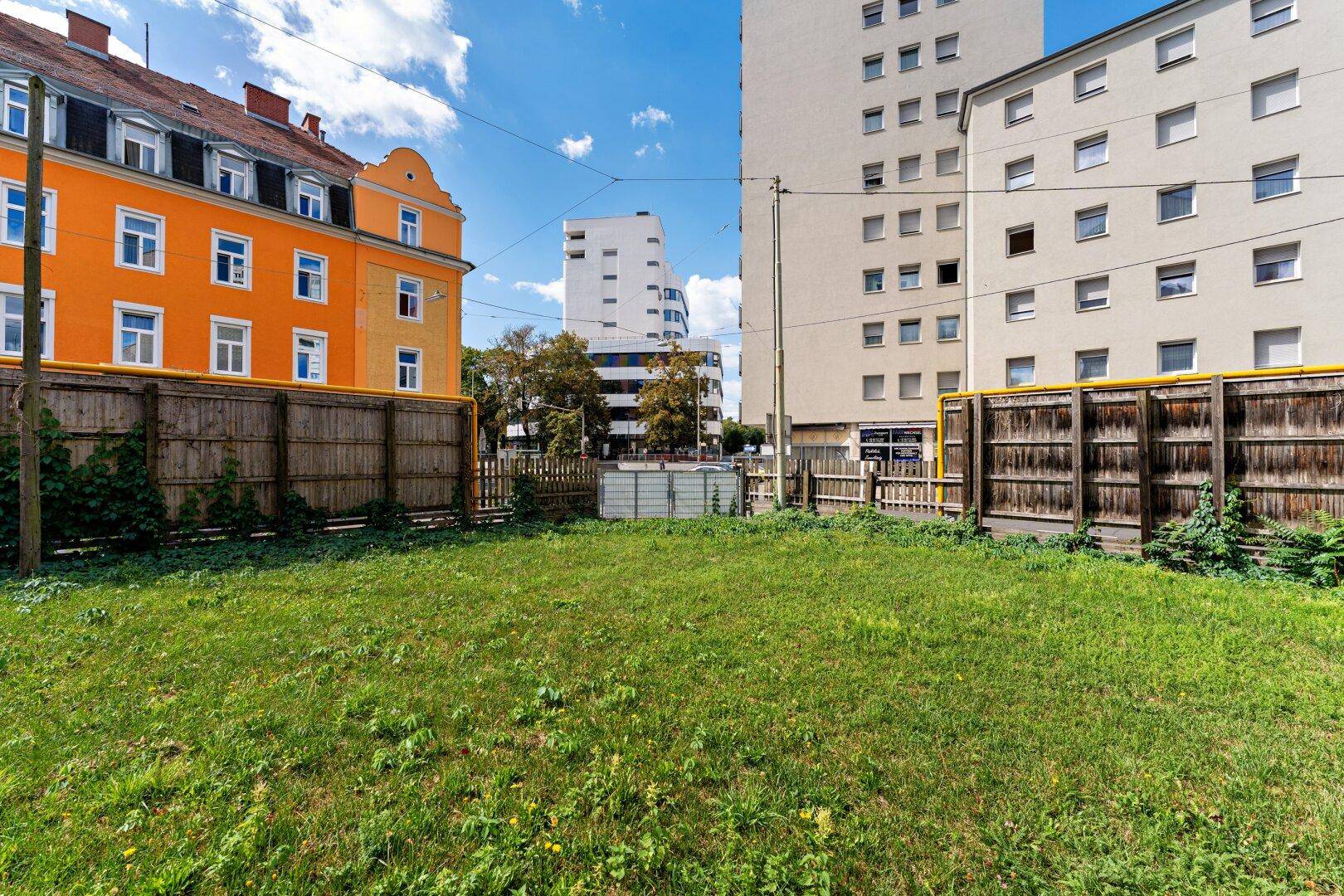 Unbebautes Grundstück mit grüner Grasfläche, umgeben von Holzzäunen und städtischen Gebäuden.