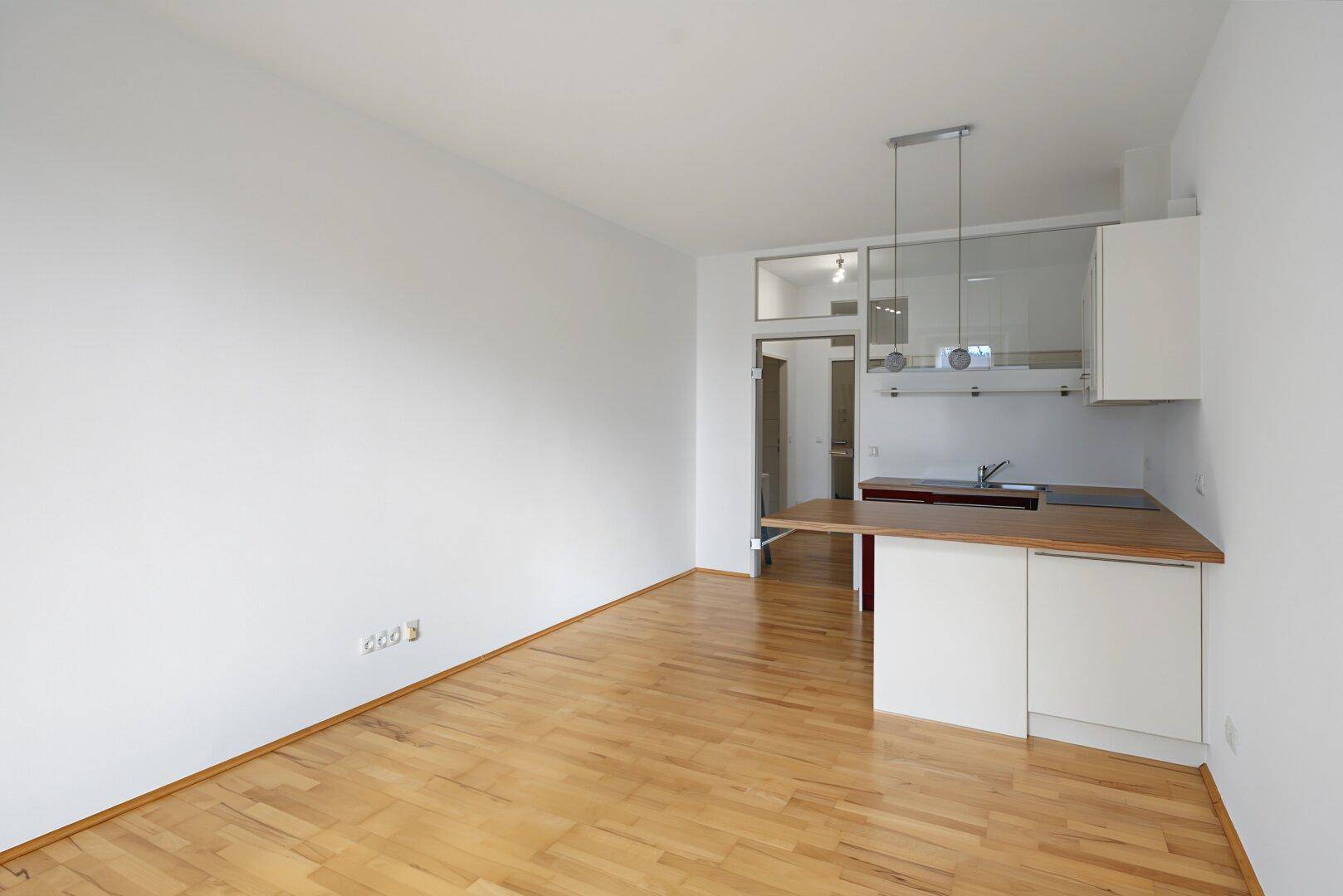 Heller Wohnbereich mit Holzboden und integrierter Küchenzeile im Hintergrund.