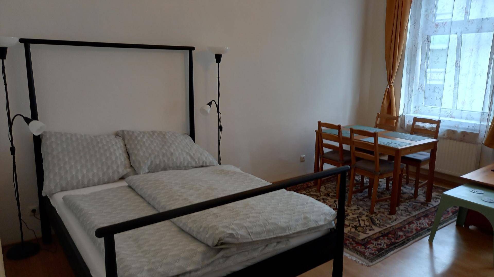 Schlafbereich mit Doppelbett und Stehlampen, angrenzend an einen Essbereich mit Holztisch.