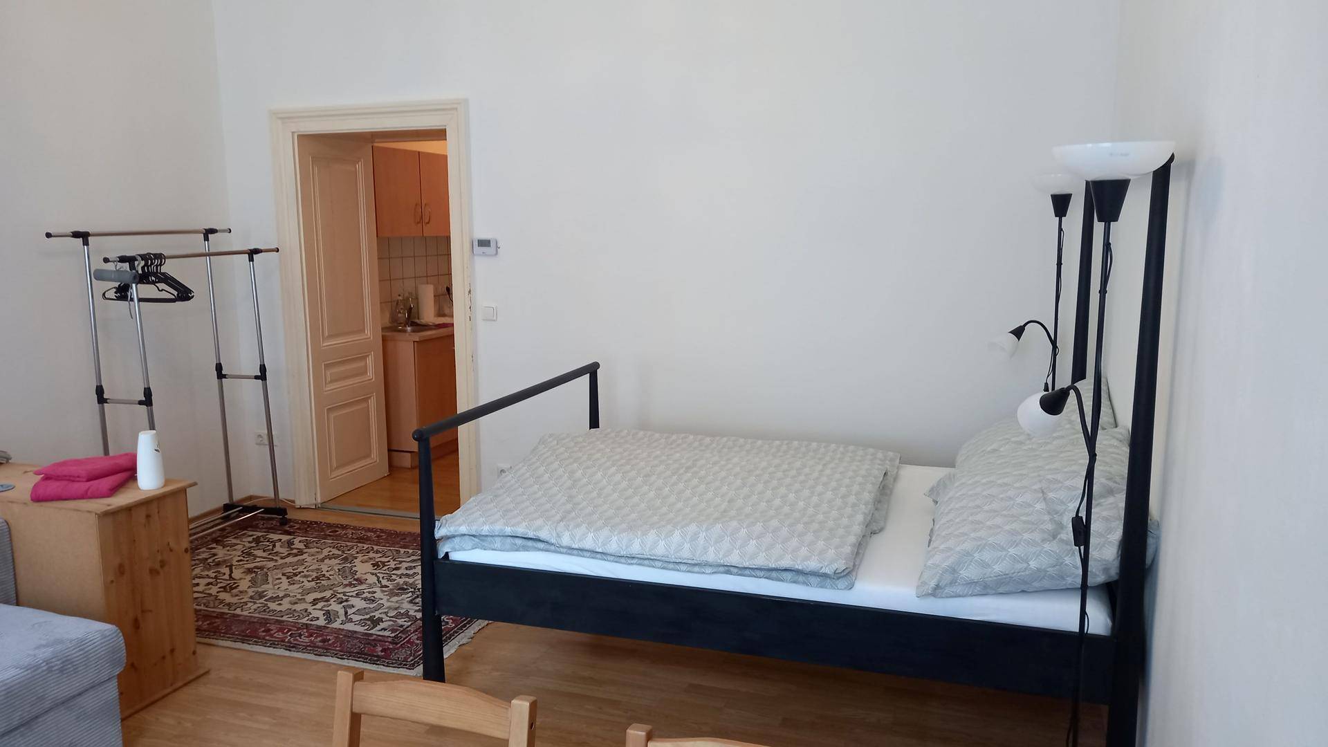 Schlafzimmer mit Bett und Kleiderständer, mit Blick durch die Tür in eine angrenzende Küche.
