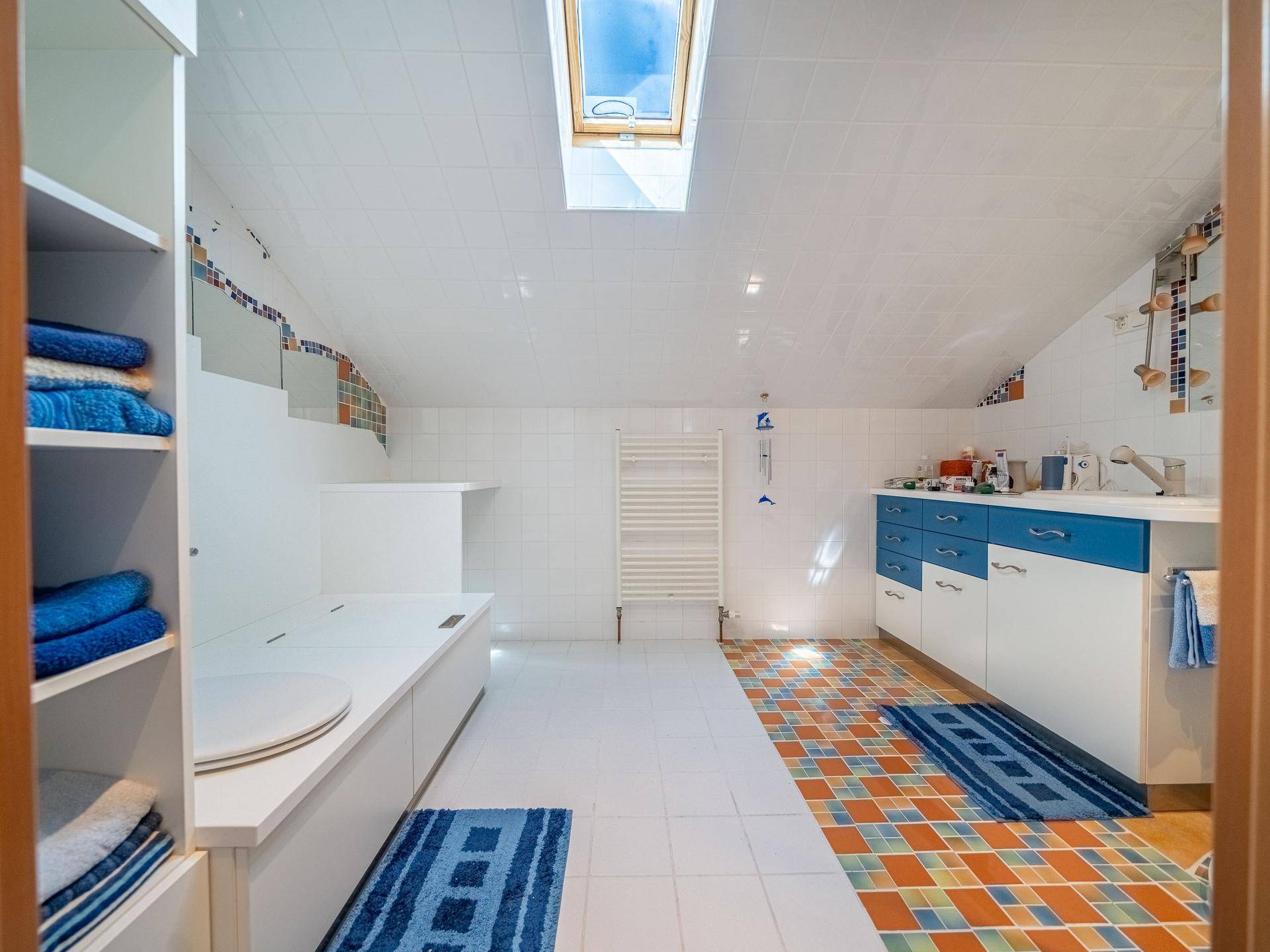 Geräumiges Badezimmer mit Dachfenster, weißen Fliesen und einem Waschtisch mit blauen Akzenten.