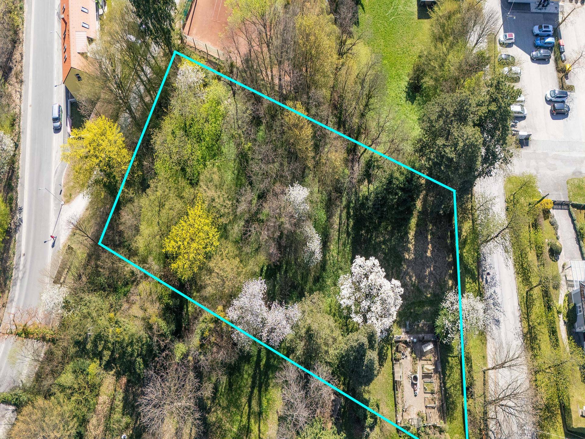 Luftaufnahme eines von Bäumen umgebenen Grundstücks mit blauer Umrandung, neben einer Straße und weiteren Gebäuden.