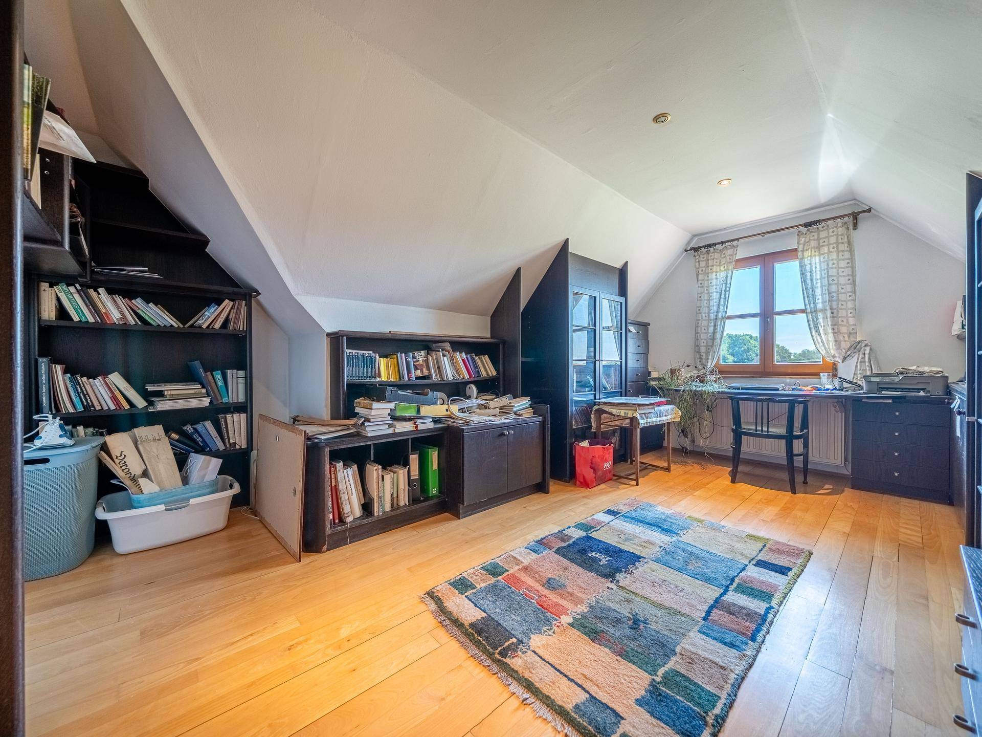 Geräumiges Dachbüro mit Holzfußboden, vielen Bücherregalen und einem Fenster mit Ausblick.