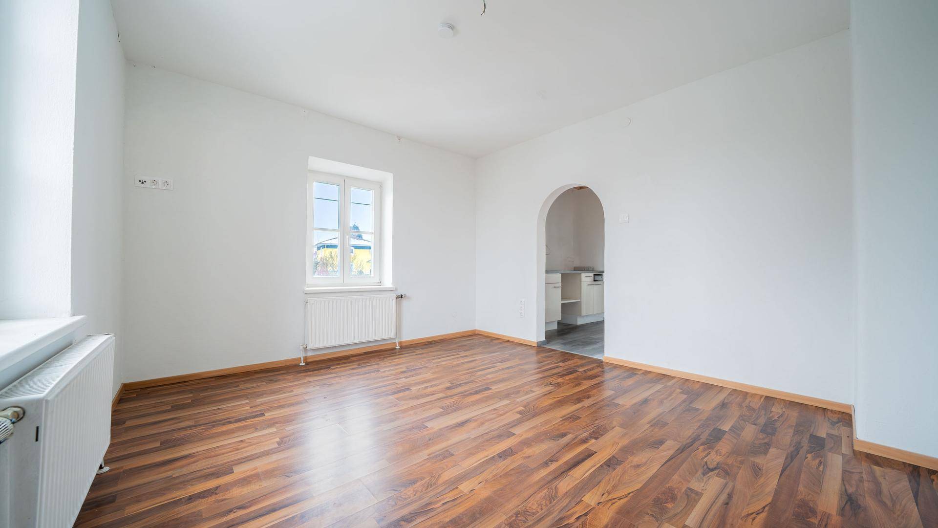 Helles, leeres Wohnzimmer mit Laminatboden, weißen Wänden und einem Fenster mit Heizkörper.