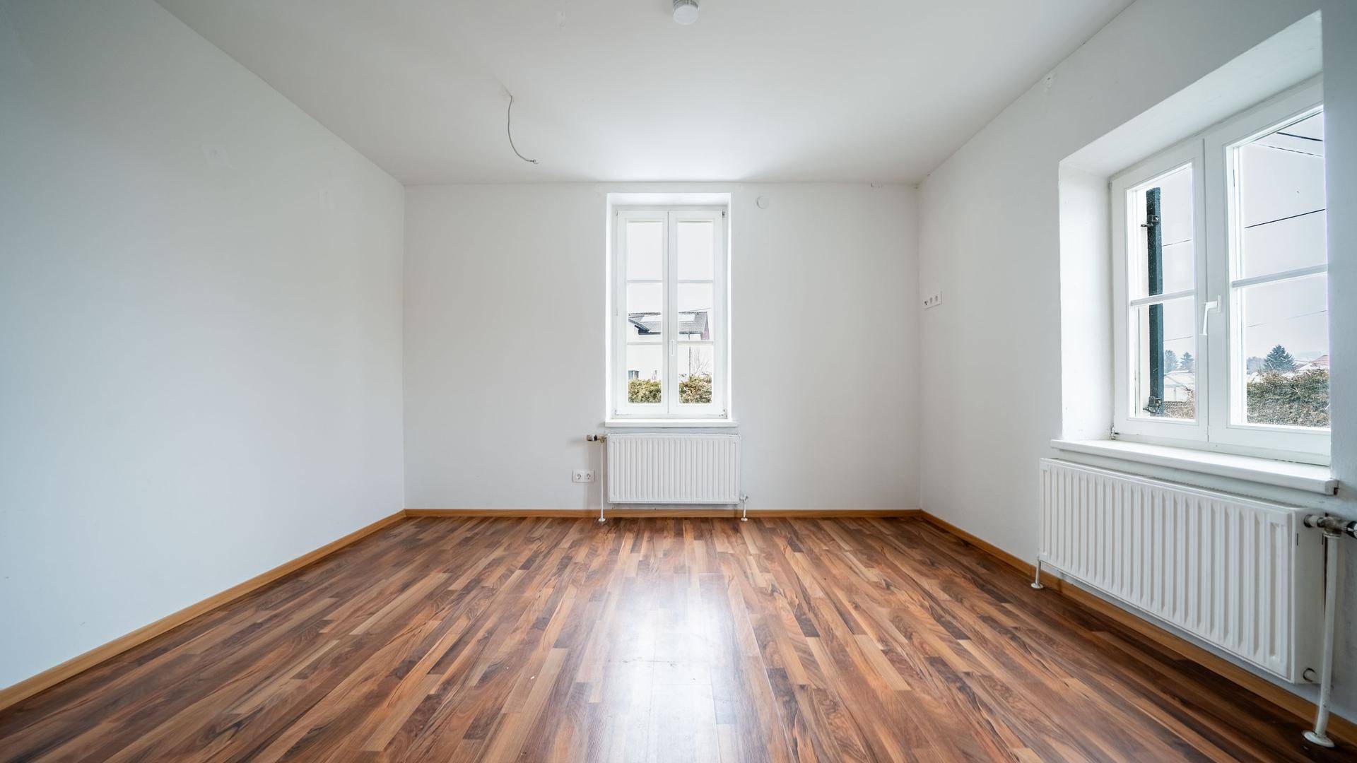 Geräumiges Wohnzimmer mit zwei Fenstern, Laminatboden und weißen Wänden, bereit zur Einrichtung.