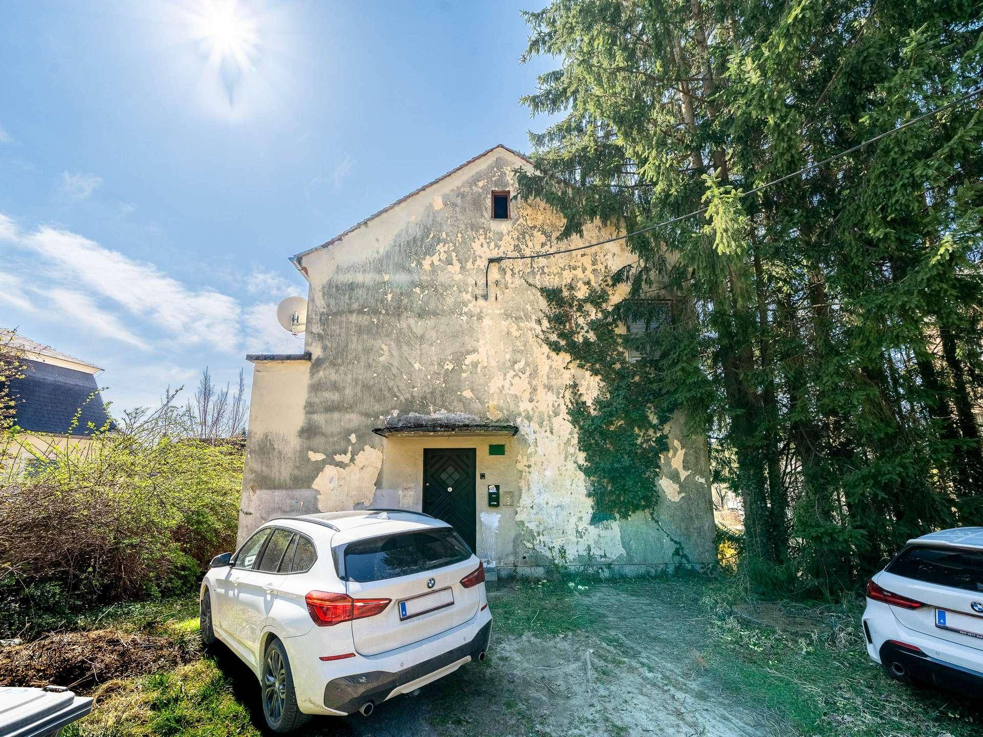 Altes Gebäude mit verputzter Fassade und zwei Autos davor, umgeben von Bäumen.