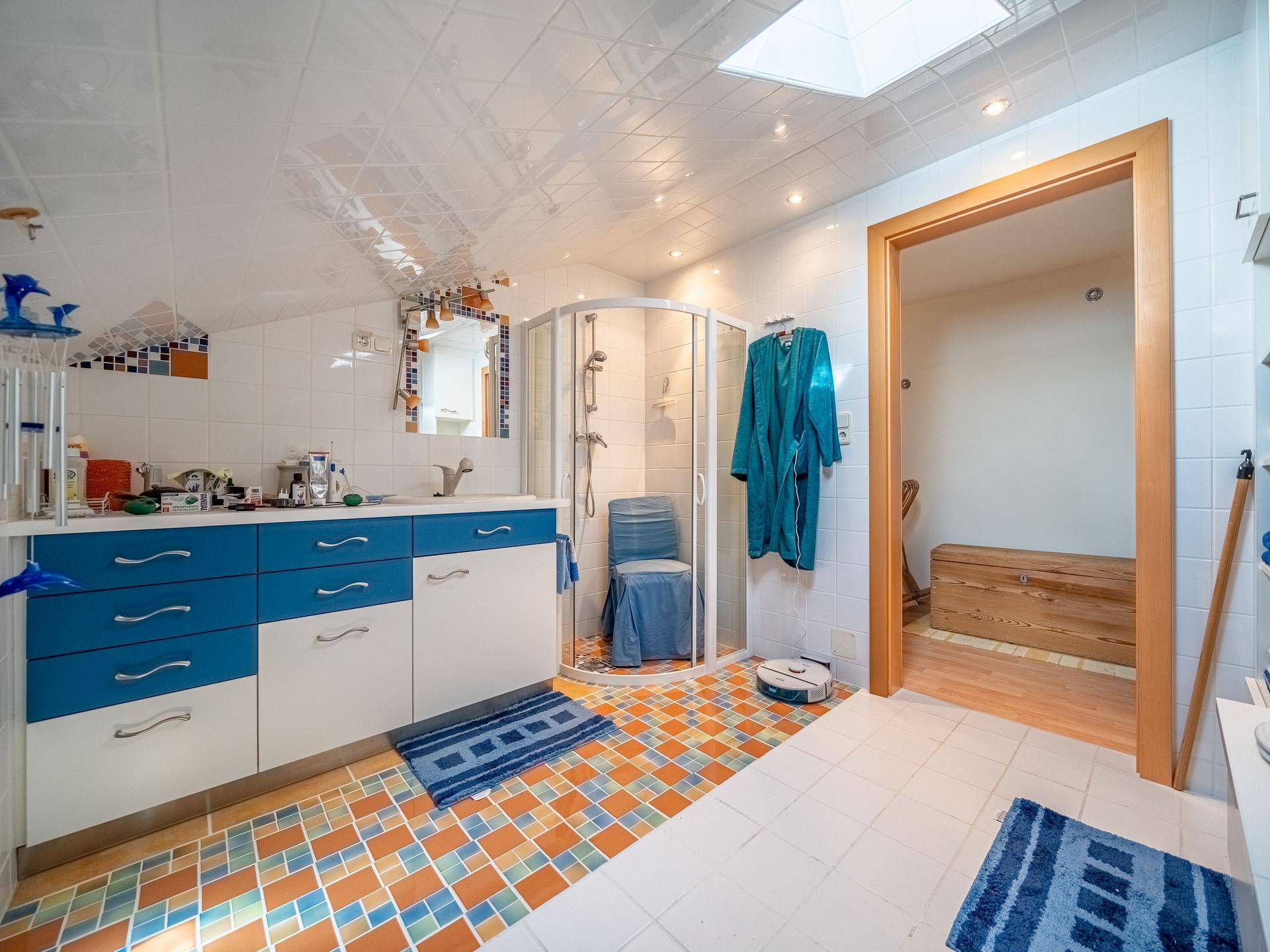 Badezimmer mit Duschkabine, blauen Schränken, farbigen Fliesen und einem Dachfenster.