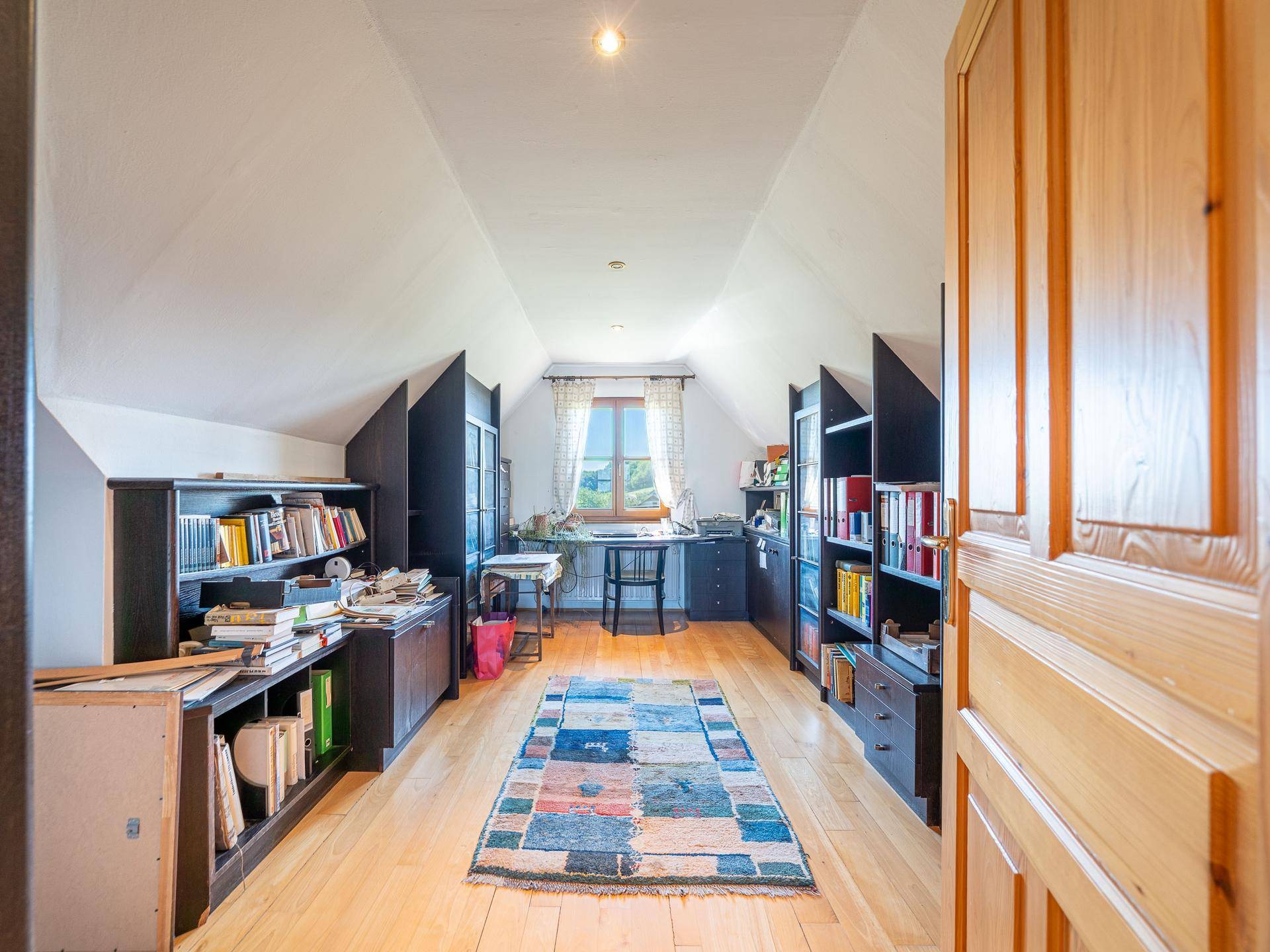 Helles Dachzimmer, eingerichtet als Büro, mit Holzfußboden, Bücherregalen und schrägen Decken.
