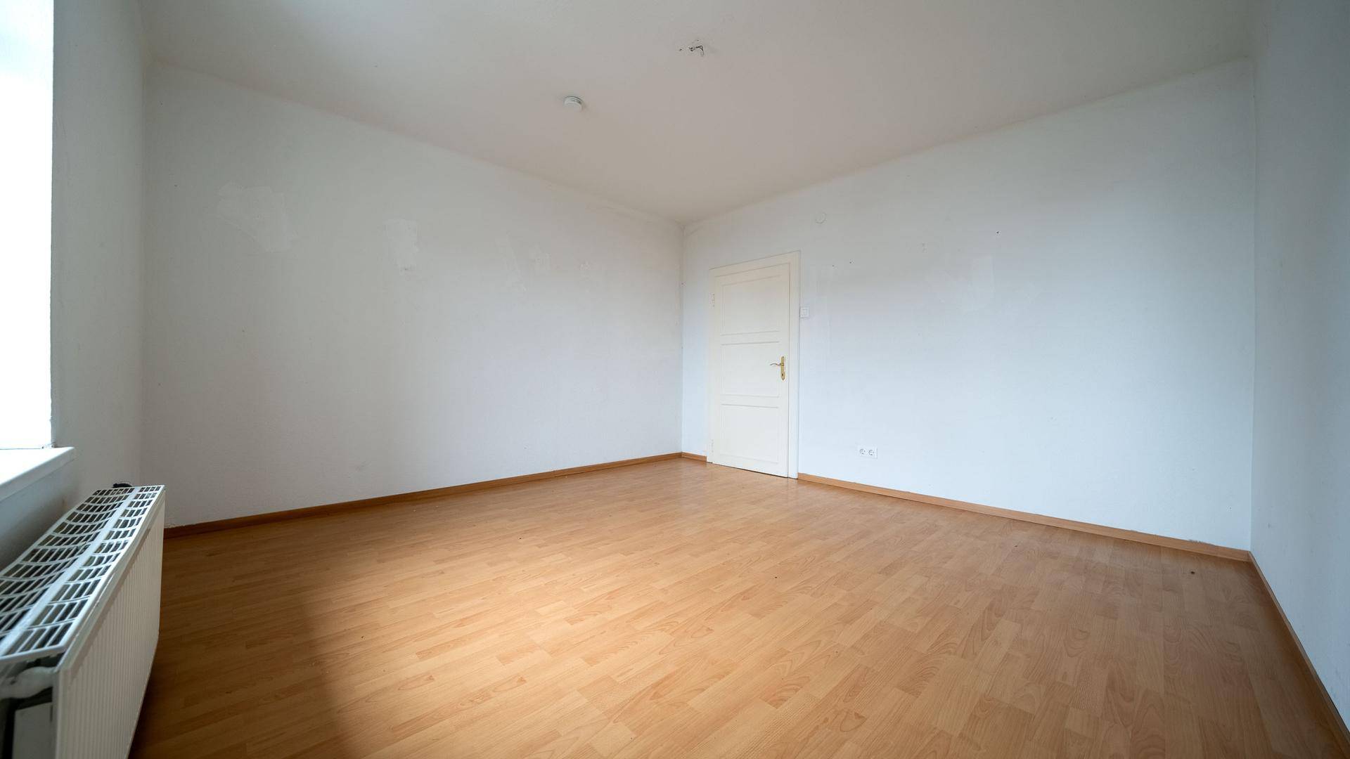 Leeres Zimmer mit Holzboden, weißen Wänden und einer einfachen Tür, bereit zur individuellen Gestaltung.