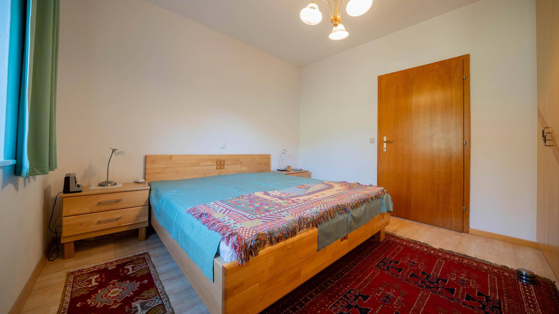 Schlafzimmer mit Holzbett, Teppich und Fensterblick ins Grüne.