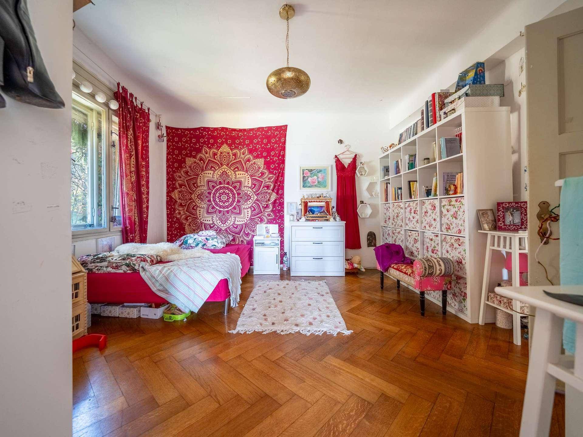 Schlafzimmer mit Parkettboden, rotem Wandteppich und Bücherregal, das eine gemütliche Atmosphäre schafft.