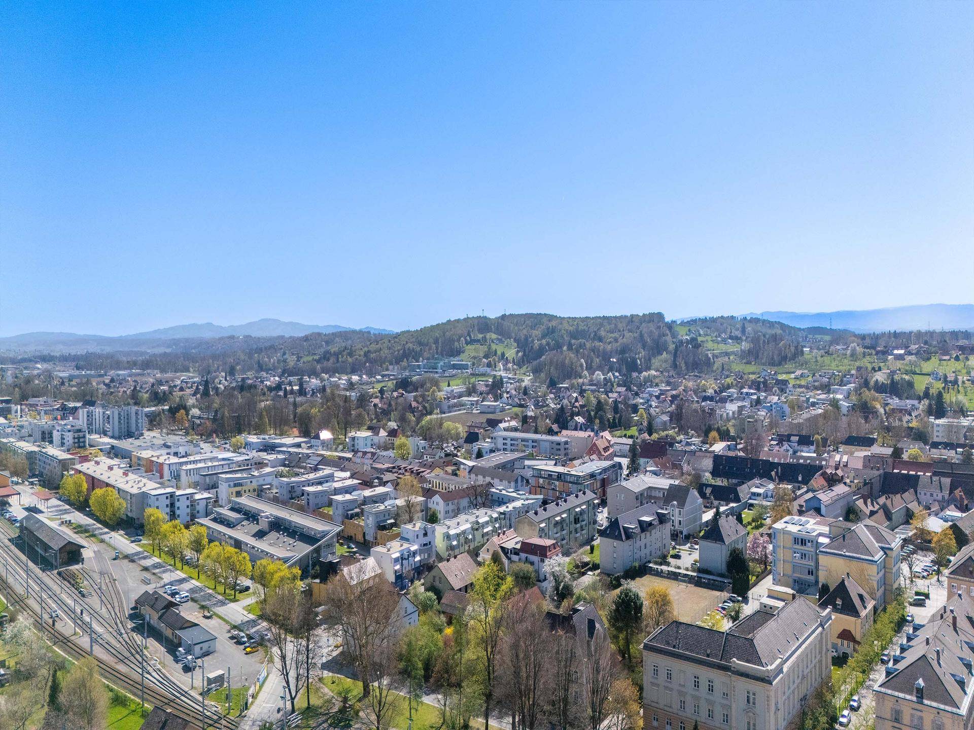 Weitläufige Luftaufnahme einer städtischen Siedlung mit vielen Gebäuden, Grünflächen und Bahngleisen.