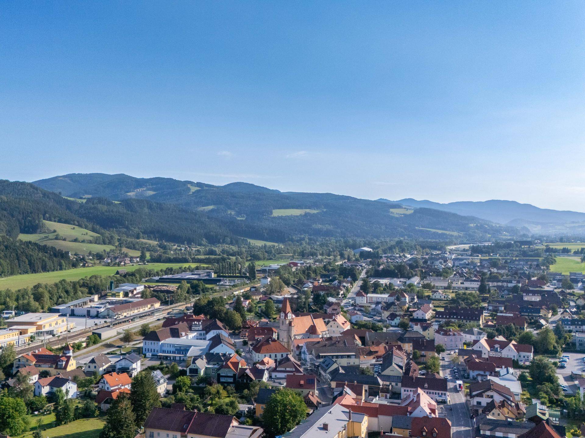 Weitläufige Luftaufnahme einer charmanten Stadt mit Kirchturm, eingebettet in eine grüne Berglandschaft.