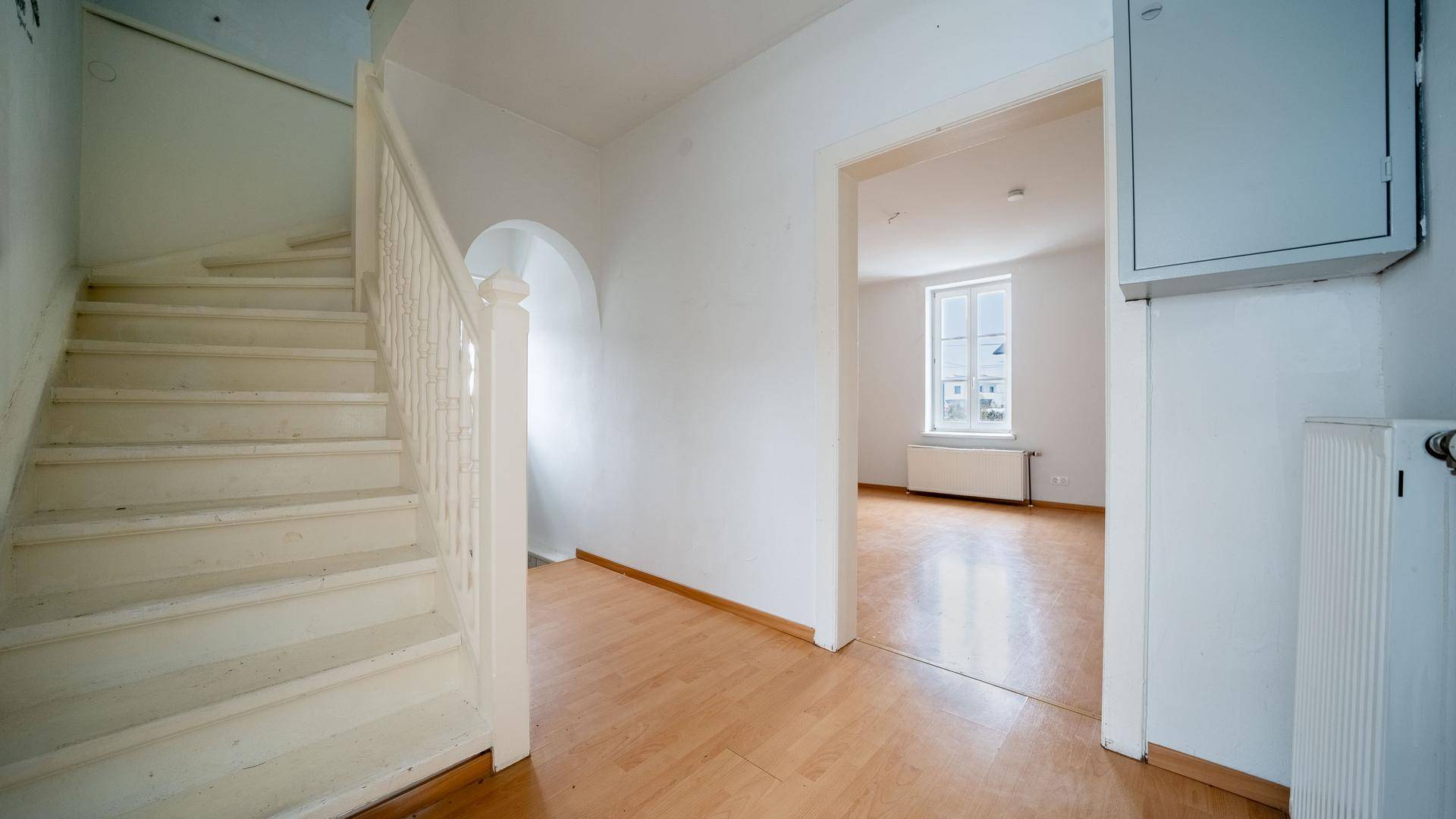 Heller Flur mit weißer Holztreppe und Zugang zu einem weiteren Zimmer, mit hellem Holzboden.