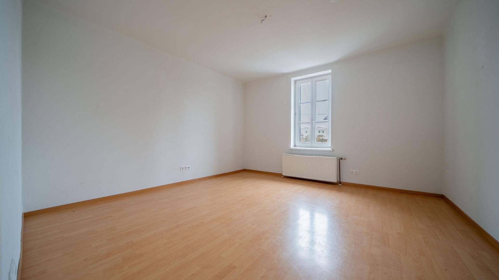 Leeres Zimmer mit hellem Holzboden, weißen Wänden und einem Fenster mit Heizkörper.