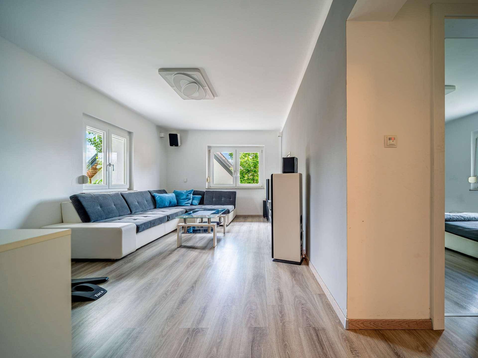 Helles Wohnzimmer mit bequemer Couch, Laminatboden und Blick ins Schlafzimmer.