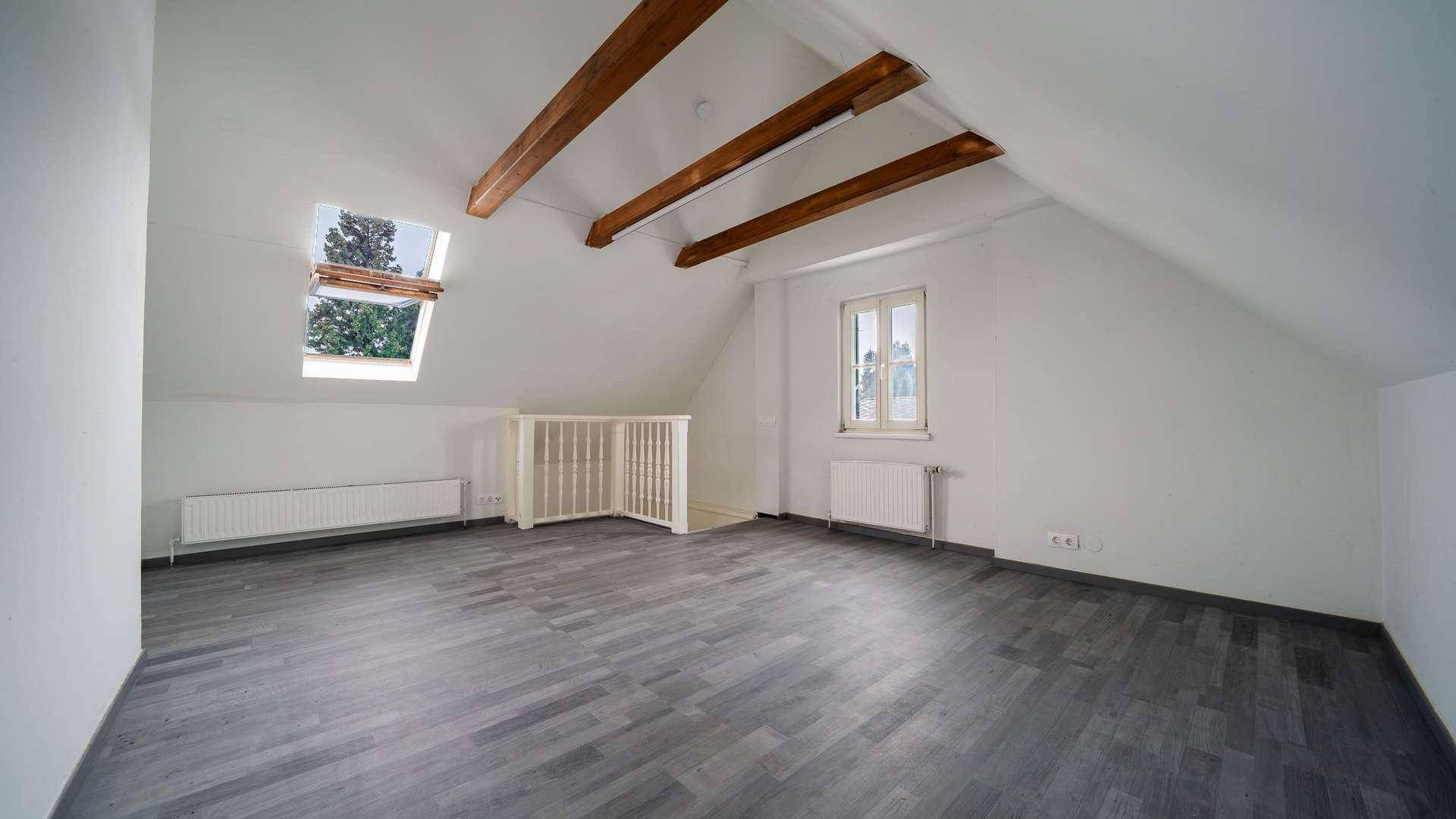 Helles Dachzimmer mit sichtbaren Holzbalken, Dachfenster und grauem Laminatboden.
