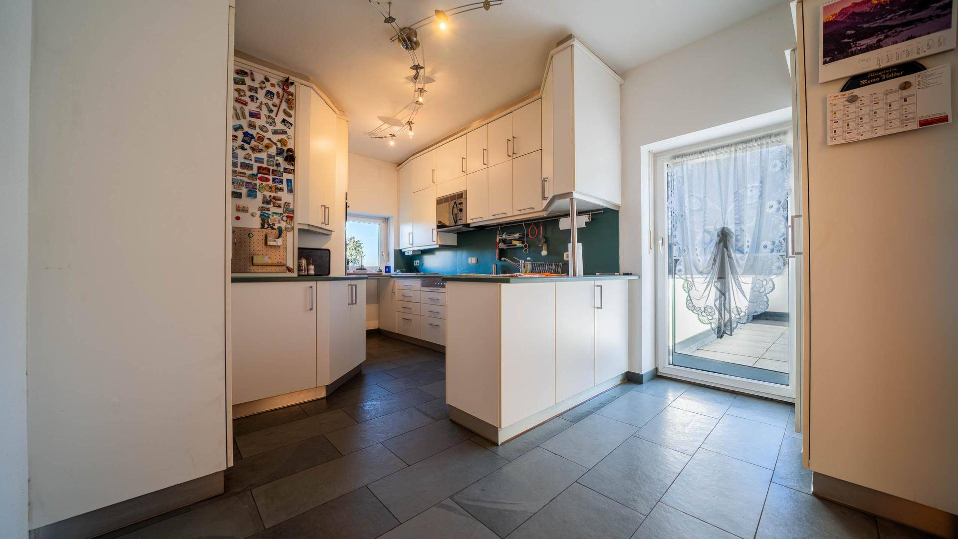 Küche mit weißen Schränken, dunklem Fliesenboden und Zugang zu einem Balkon.