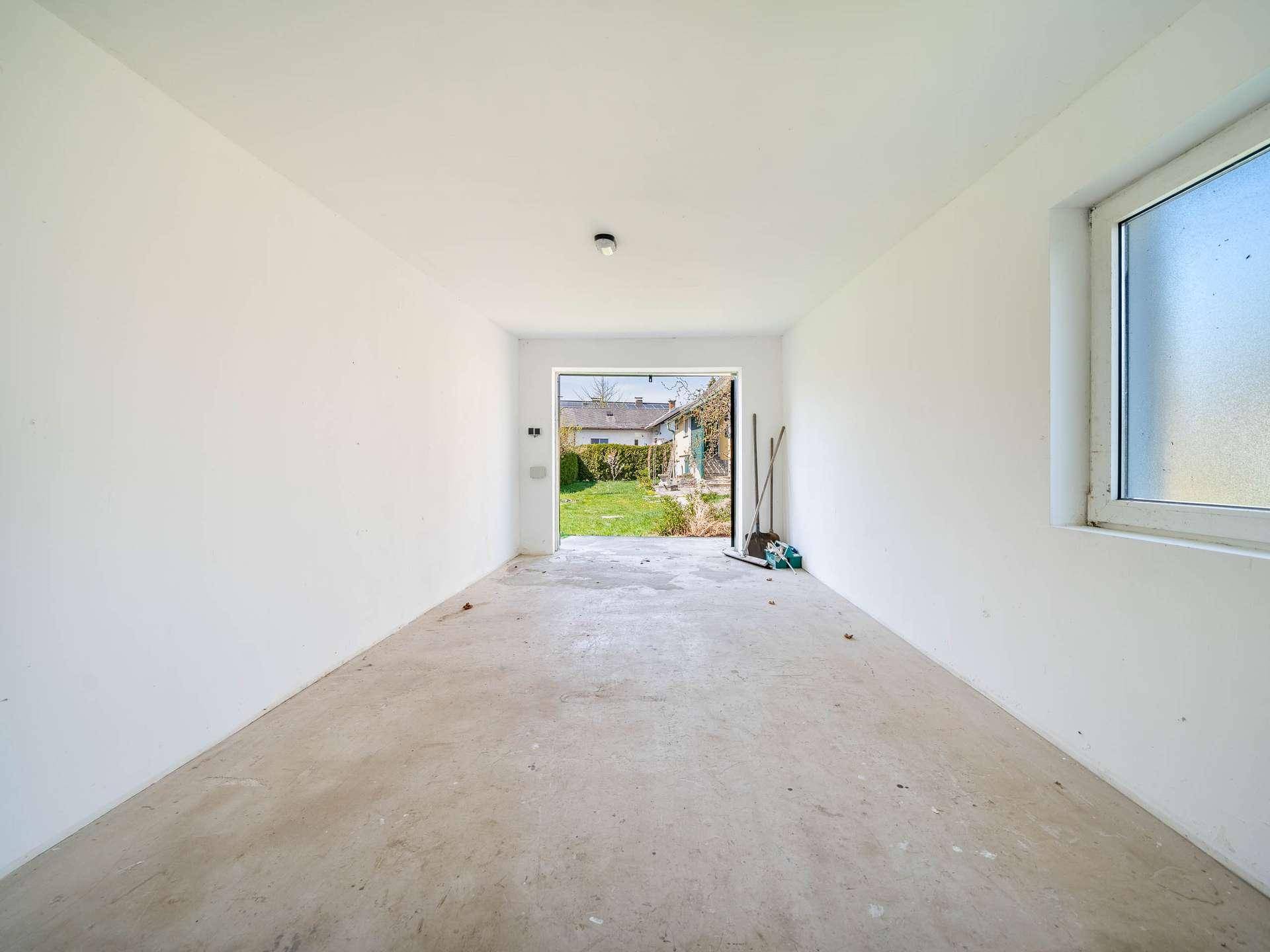Leerer Innenraum der Garage mit weißen Wänden und Betonboden, Blick in den Garten.