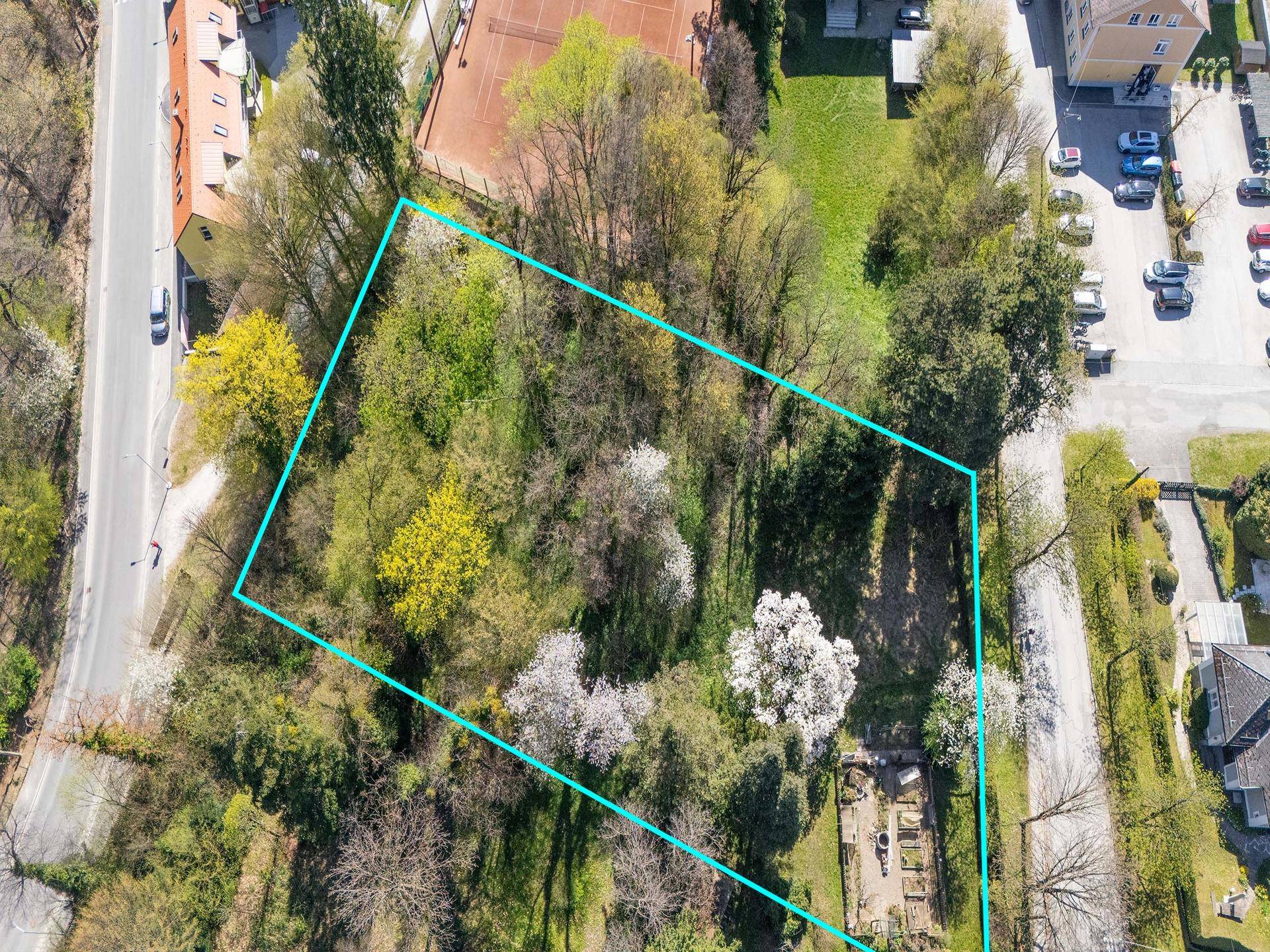 Luftaufnahme eines von Bäumen umgebenen Grundstücks mit blauer Umrandung, neben einer Straße und weiteren Gebäuden.
