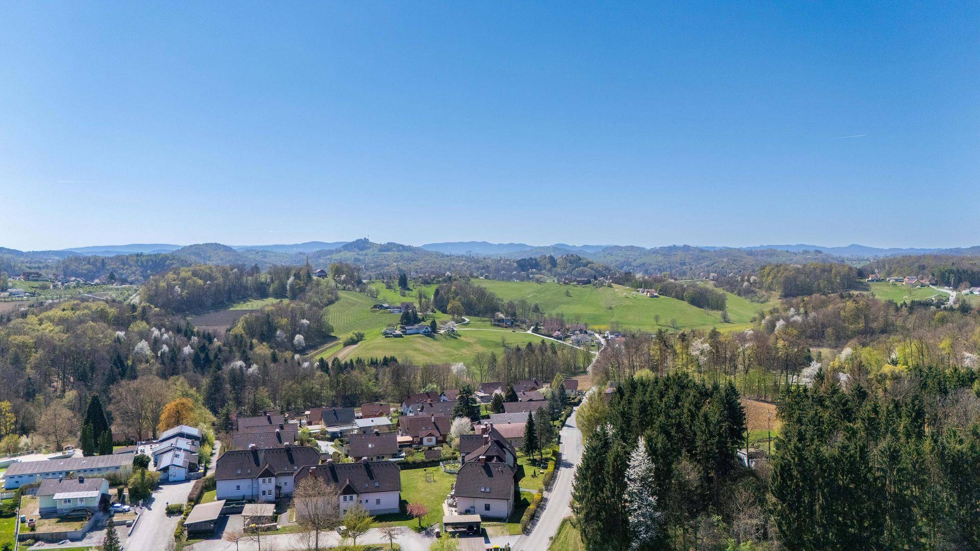 Luftaufnahme einer ländlichen Siedlung mit Häusern, Straßen und weiten grünen Hügeln im Hintergrund.
