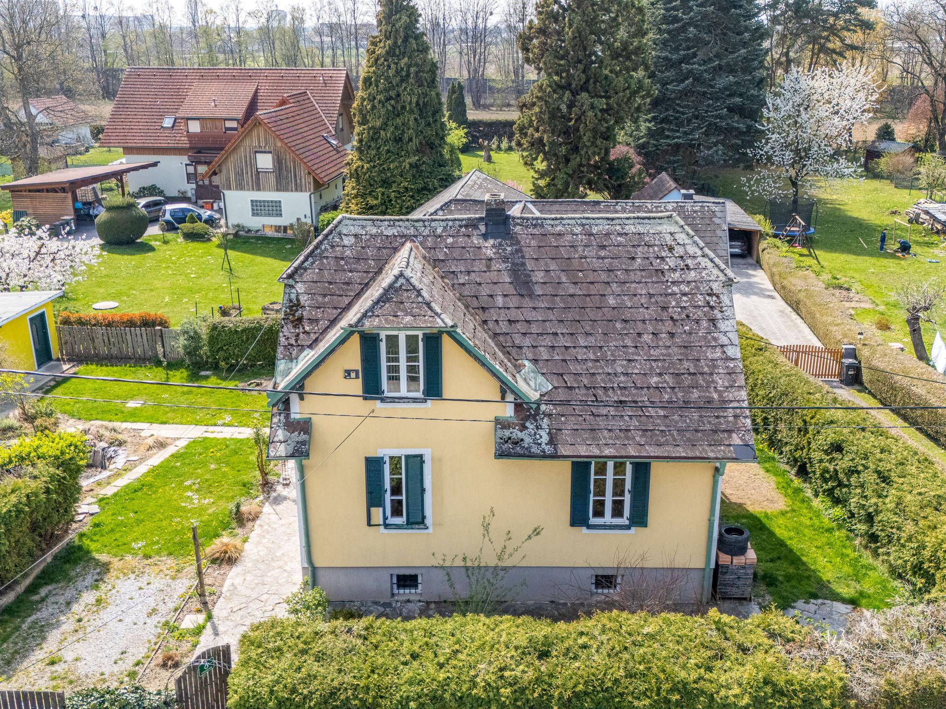 Frontale Luftaufnahme des Hauses mit gelber Fassade, umgeben von einem grünen Garten und Bäumen.