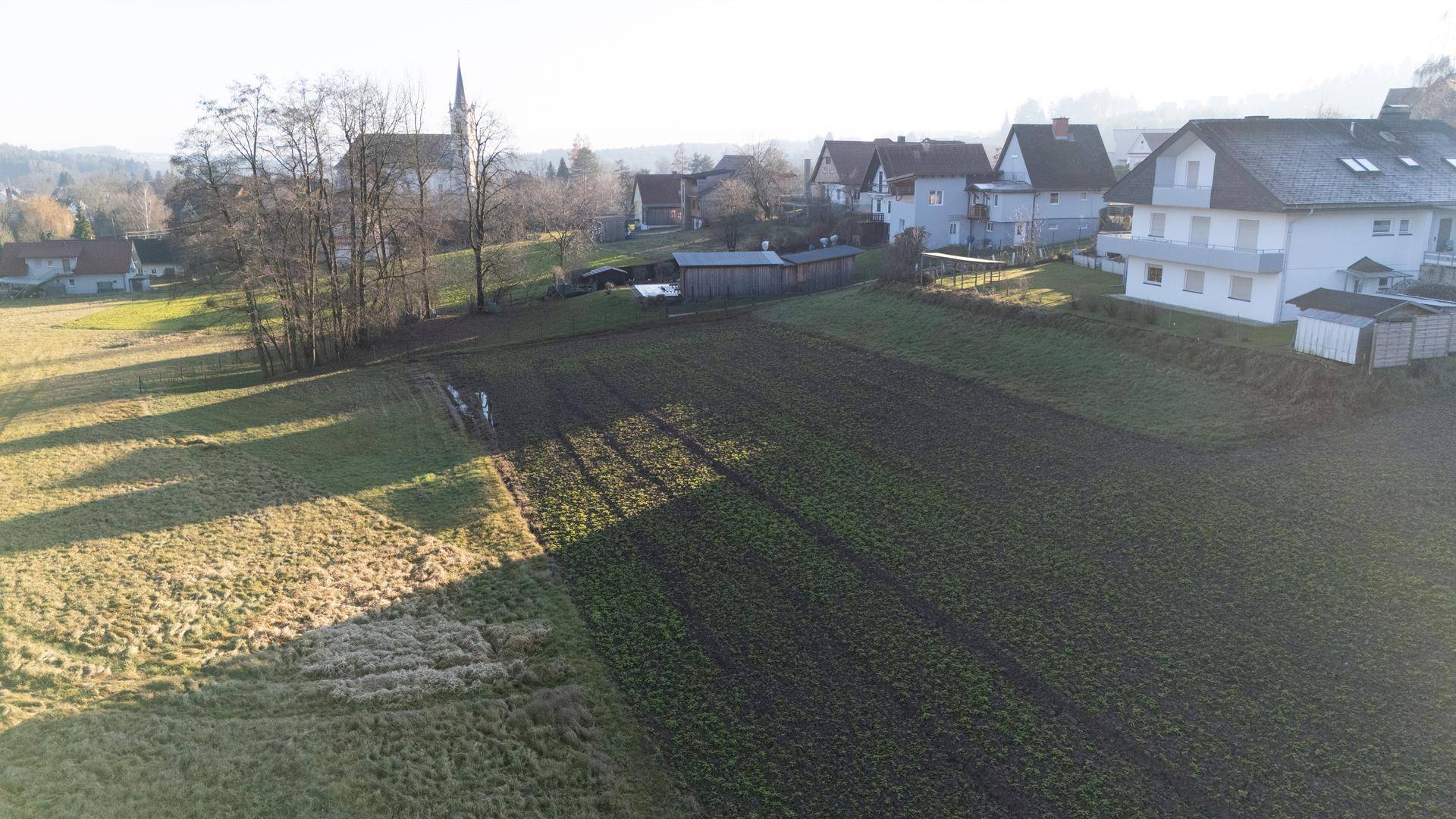 Ländliche Szenerie mit Wohnhäusern, weiten Feldern und der markanten Dorfkirche in der Ferne unter klarem Himmel.